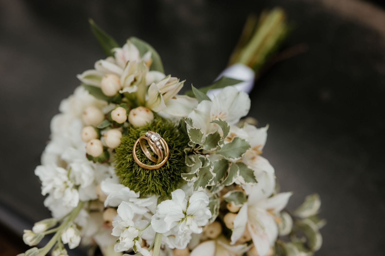 Gold wedding rings rest on a lush white bridal bouquet with green accents and delicate blooms.
