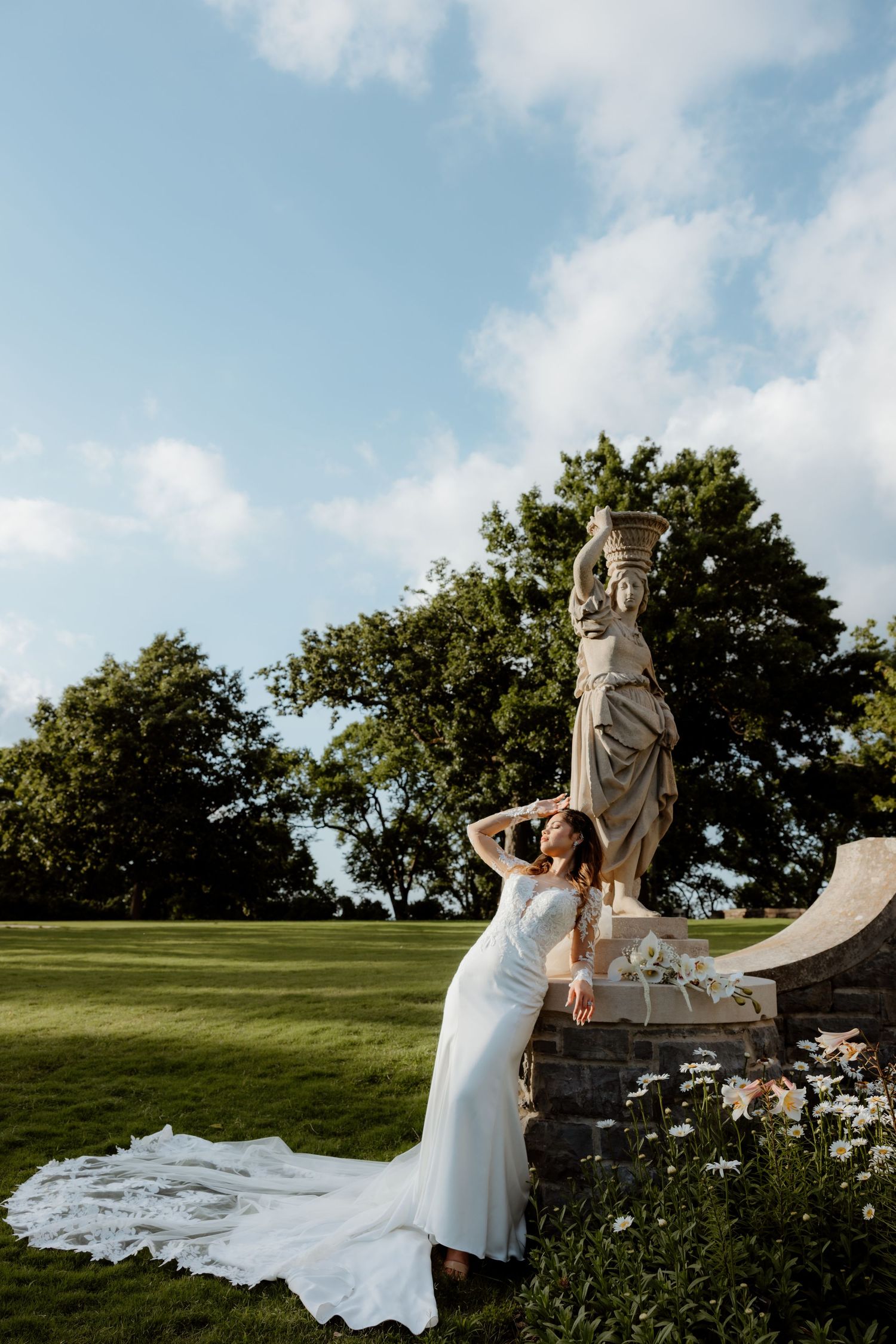 Bride in white gown with long train poses by classical stone statue in lush garden under blue sky.