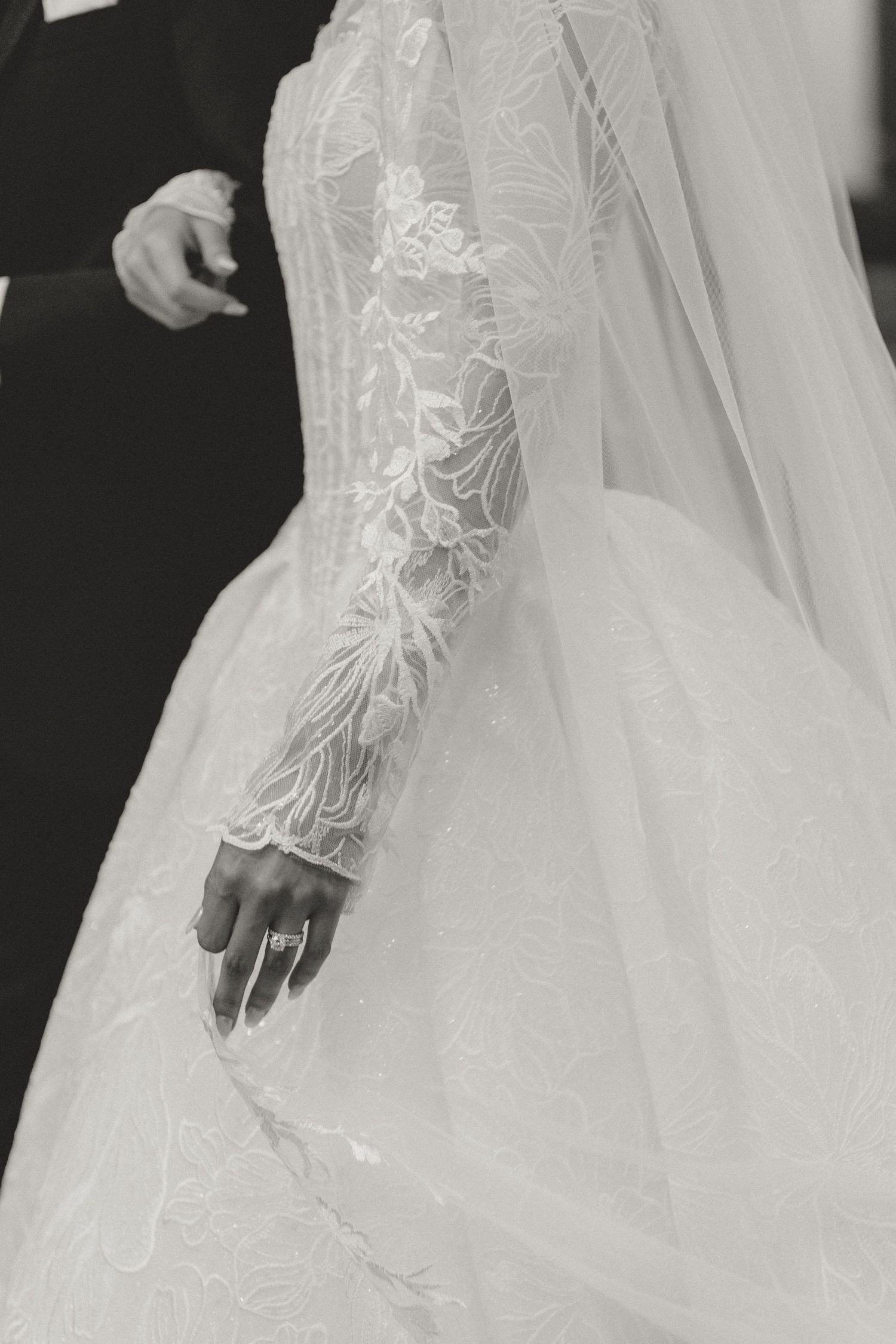 Close-up of bride's lace long-sleeve wedding gown with diamond engagement ring in black and white.