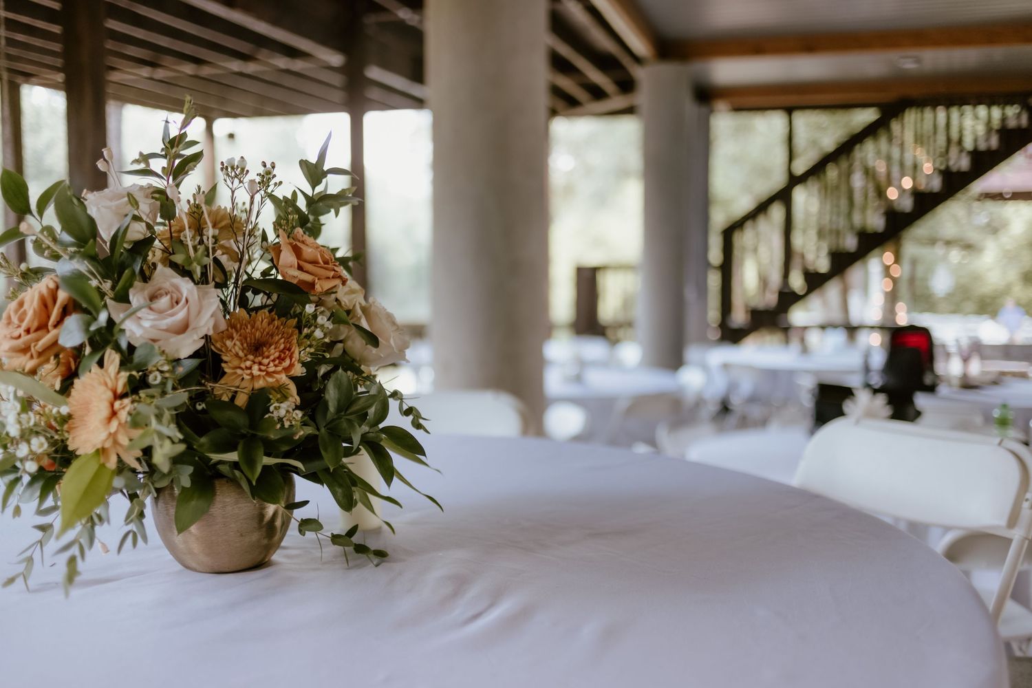 An elegant floral centerpiece with white and peach flowers sits atop a white tablecloth at a reception venue.