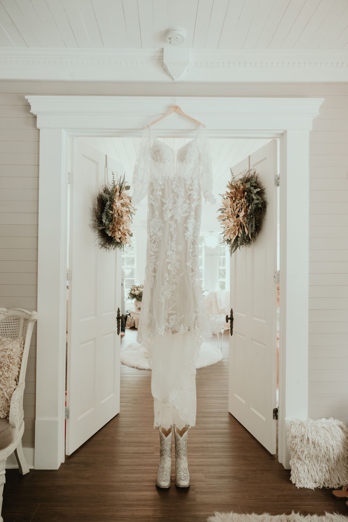A white lace wedding dress hangs between double doors adorned with dried floral wreaths and cowboy boots beneath.