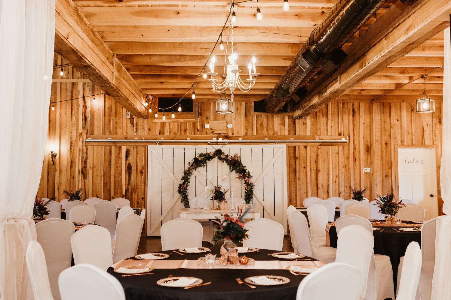 Rustic barn wedding reception venue with wooden walls, chandelier lighting, and elegant table settings dressed in white linens.
