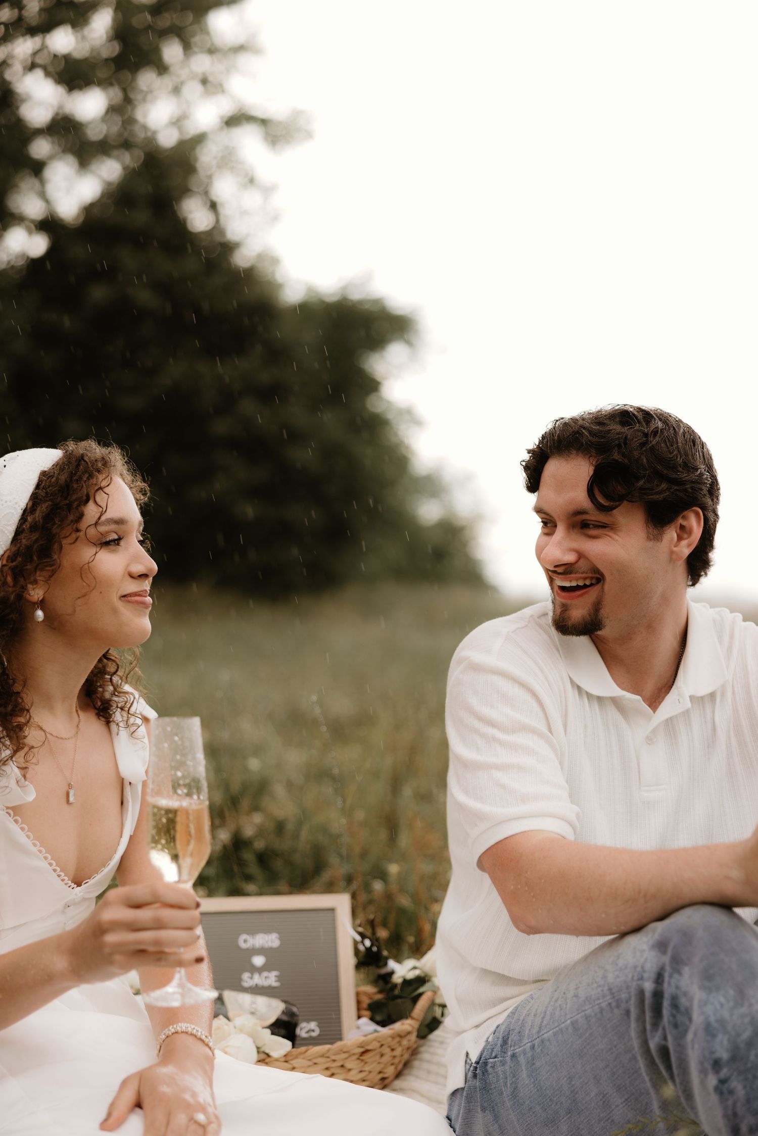 A couple having a romantic picnic outdoors with wine and laughter in soft natural lighting.