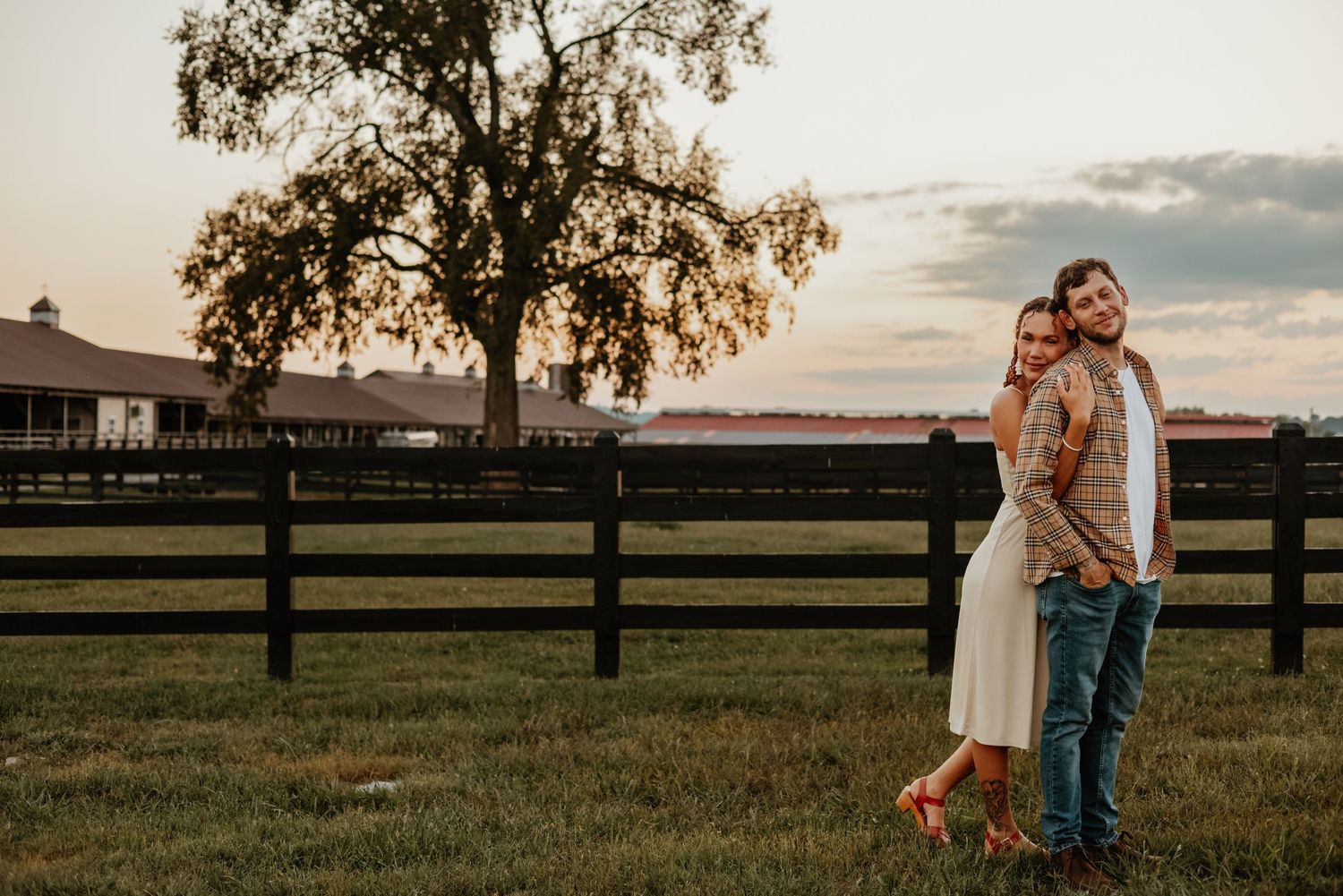 A couple stands together at sunset by a wooden fence on a rural farm with a large tree in the background.