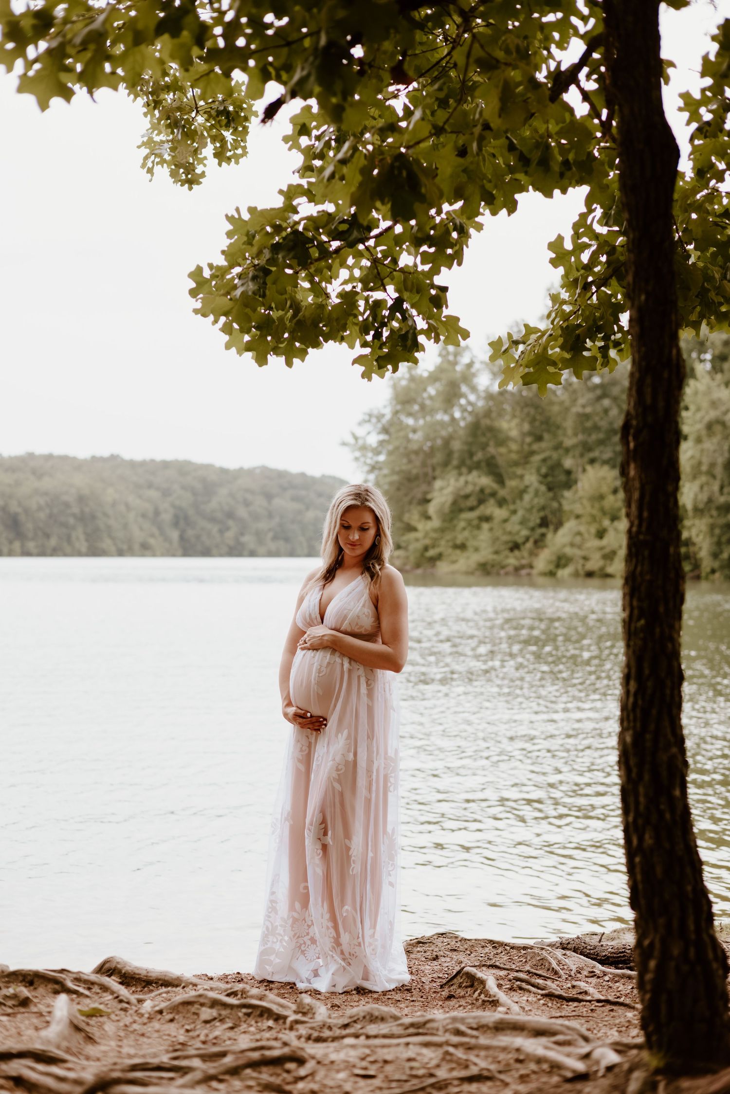A pregnant woman in a flowy white lace dress stands by a serene lake while golden sunlight filters through tree branches.