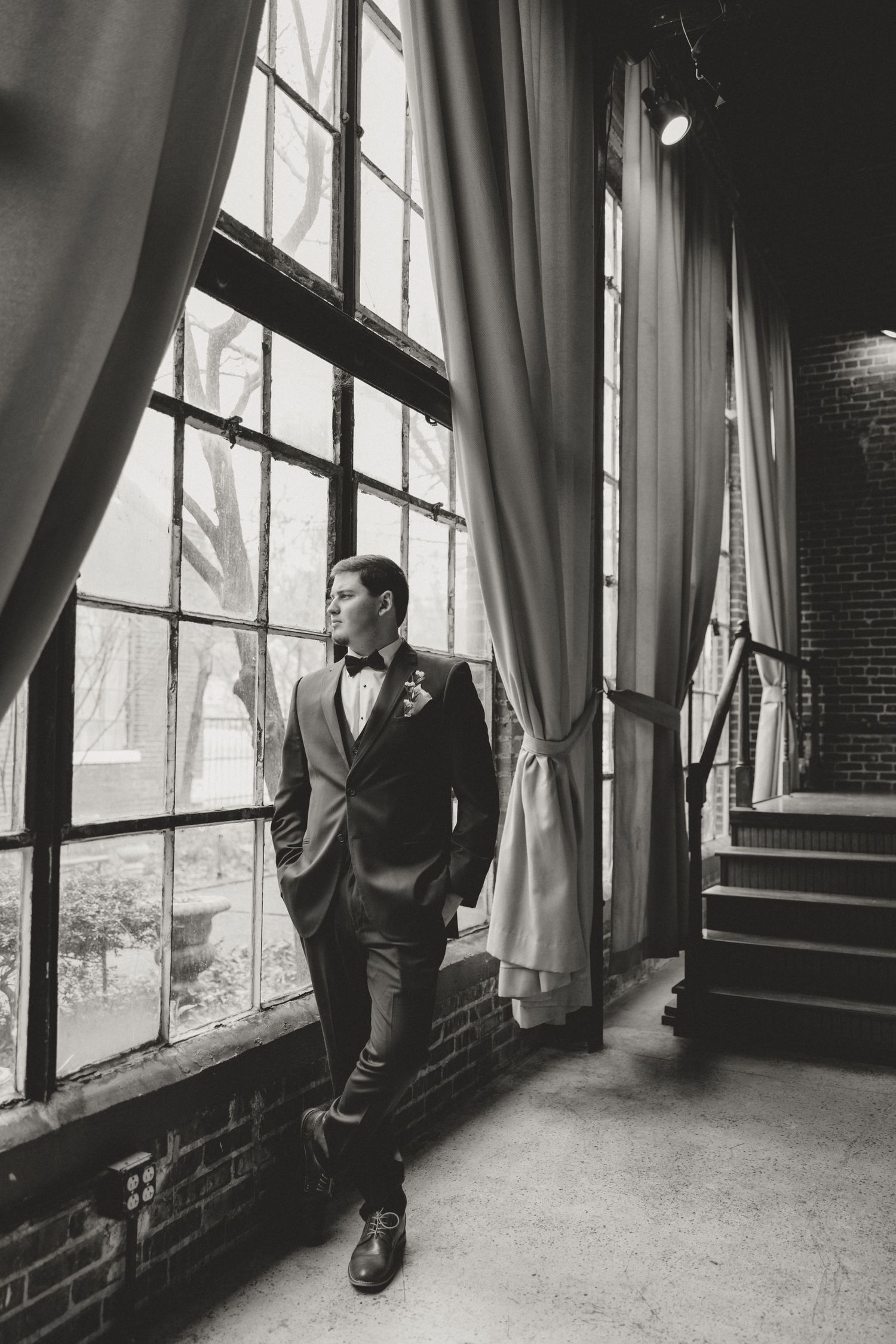 A groom in a classic tuxedo poses by tall industrial windows in a vintage brick building.