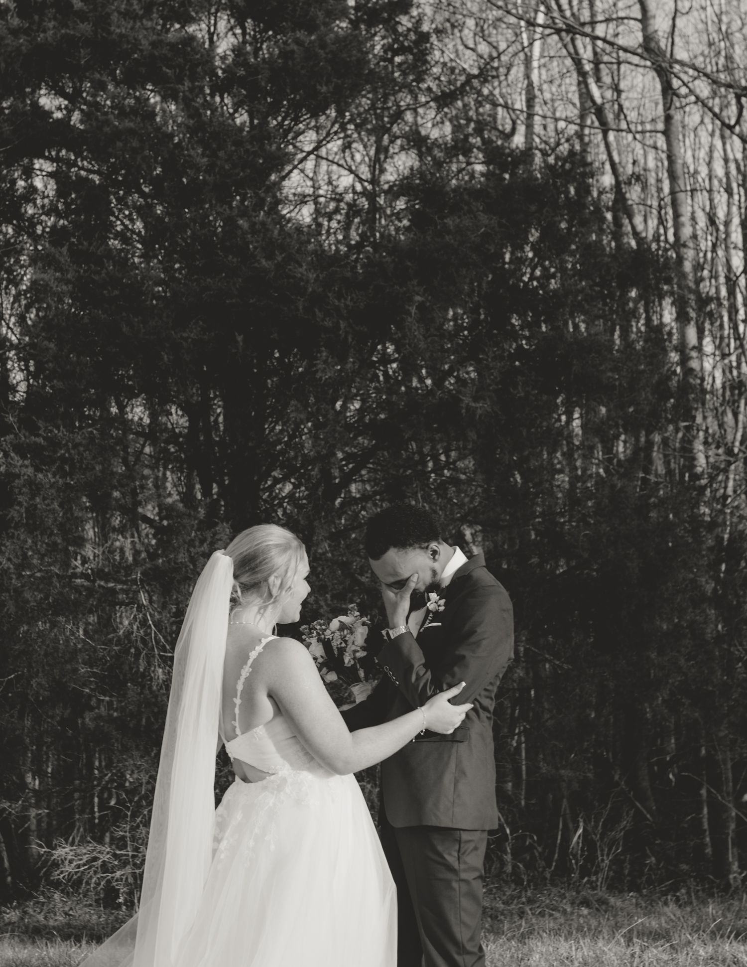 A black and white wedding photo shows a tender moment between newlyweds in a natural outdoor setting.