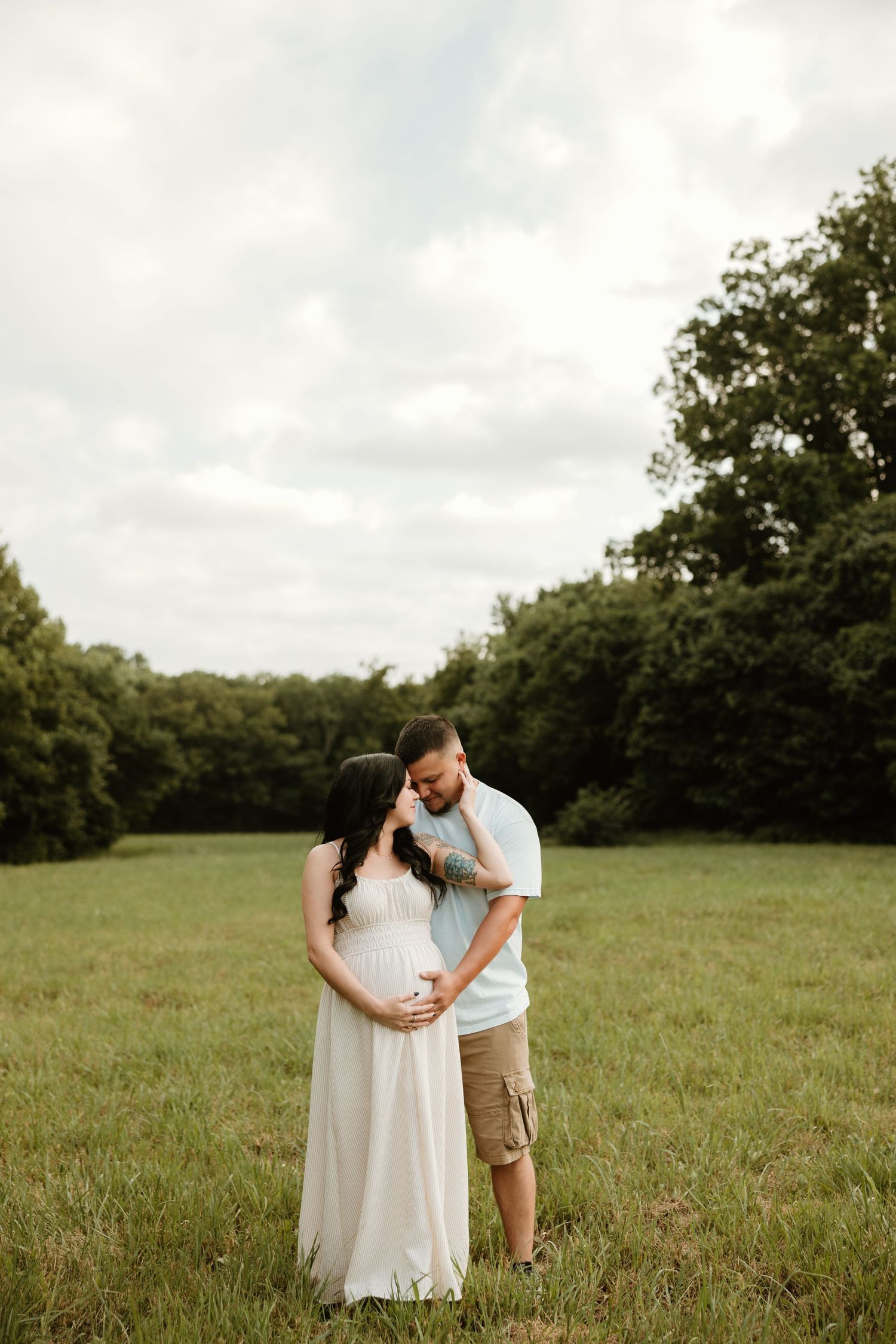 A romantic maternity photo shows a couple embracing in a grassy field with trees in the background.