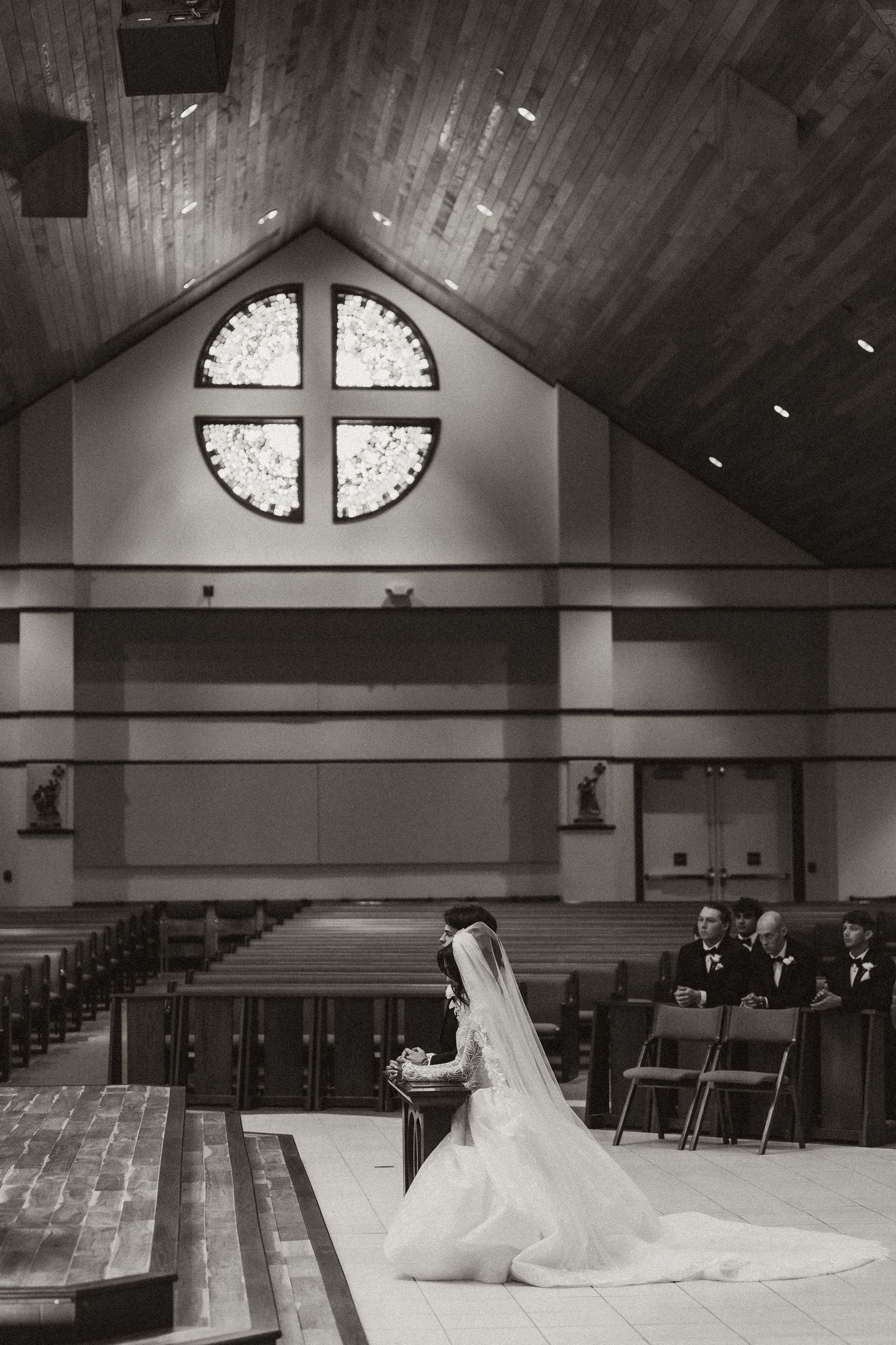 Bride kneeling at church altar in elegant white gown and veil beneath a cross stained glass window.