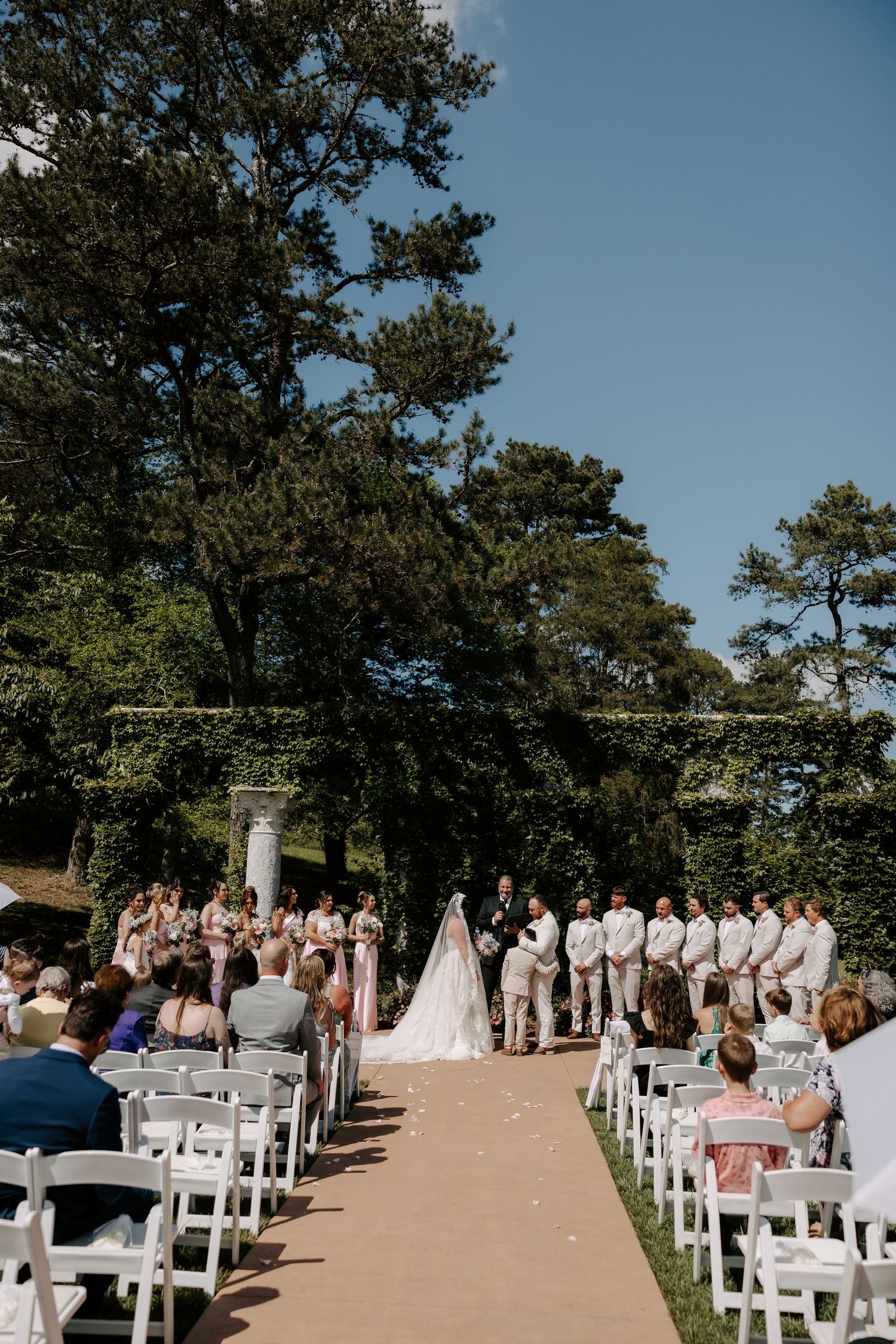 Outdoor garden wedding ceremony with bride and groom at altar, guests seated in white chairs along a petal-lined aisle.