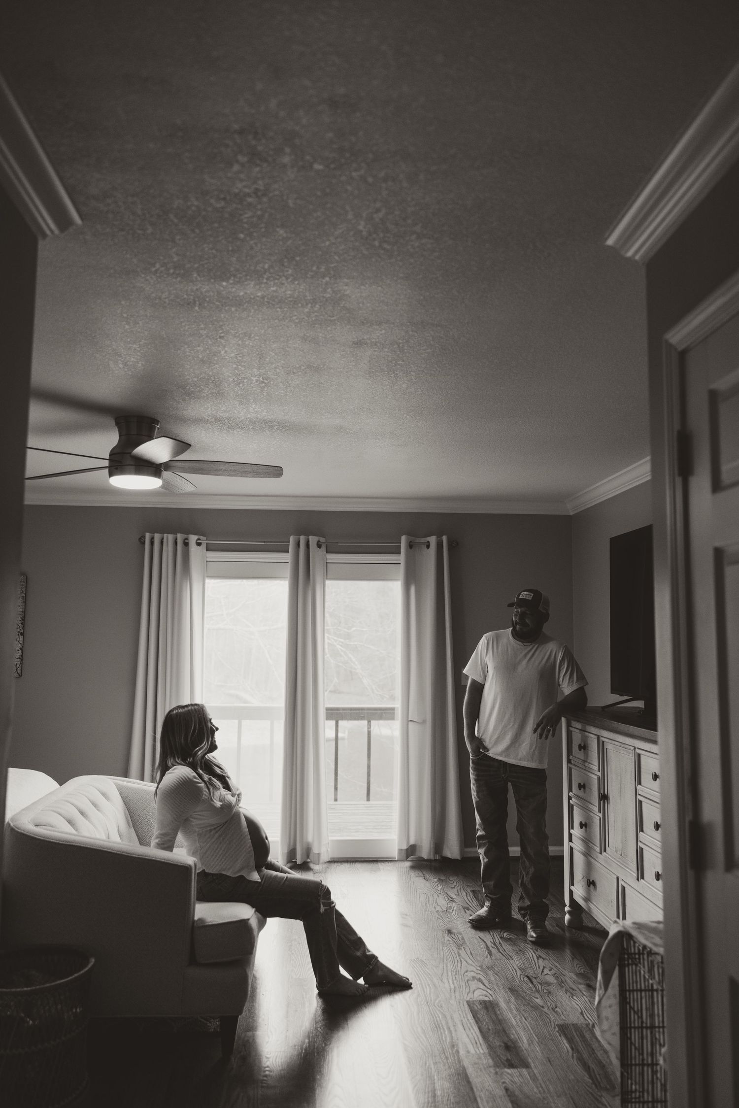 Black and white photo of a couple in a living room, woman sitting on sofa, man standing near dresser by window.