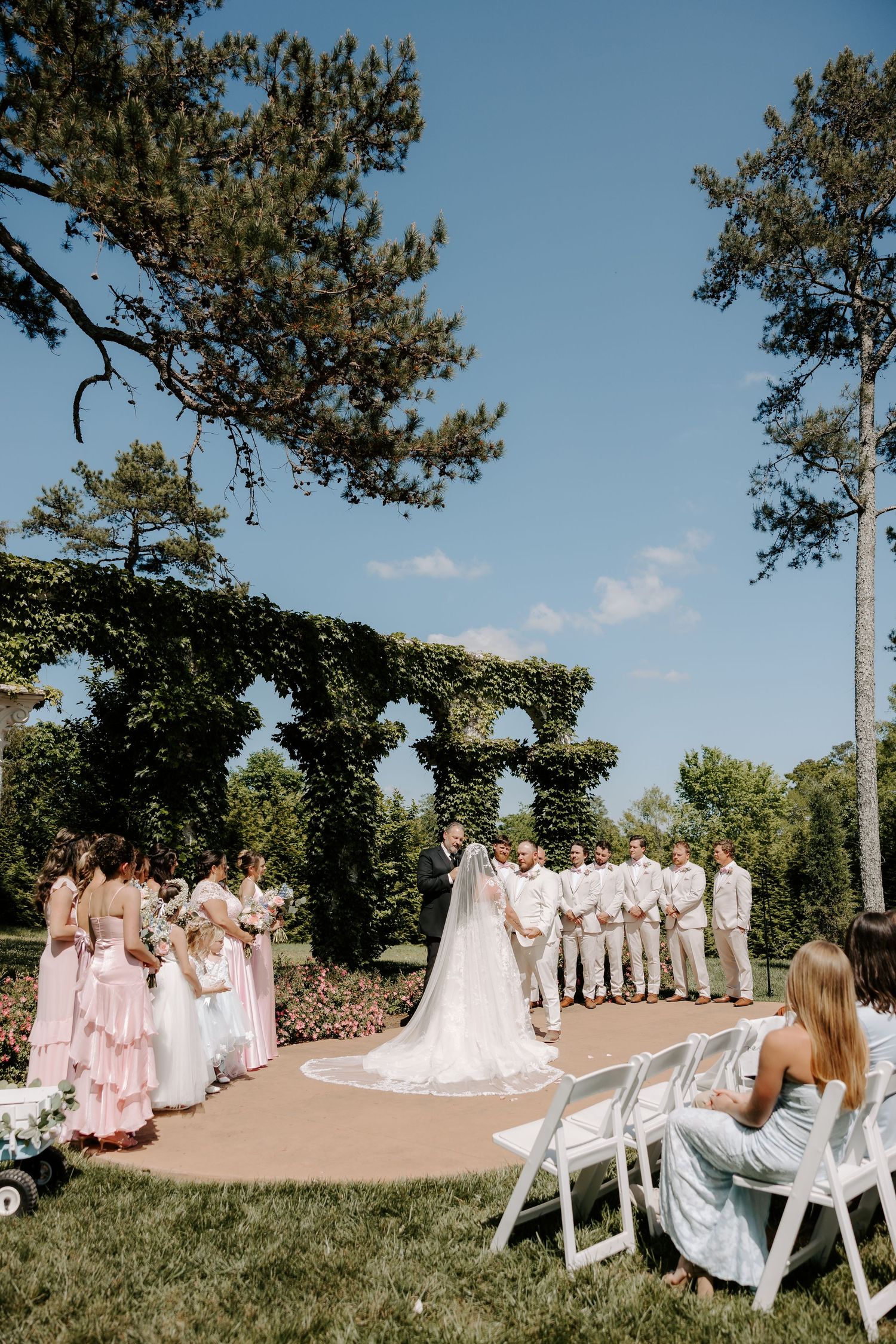 Outdoor wedding ceremony with bride and groom at altar, bridesmaids in pink, groomsmen in tan, surrounded by lush greenery.