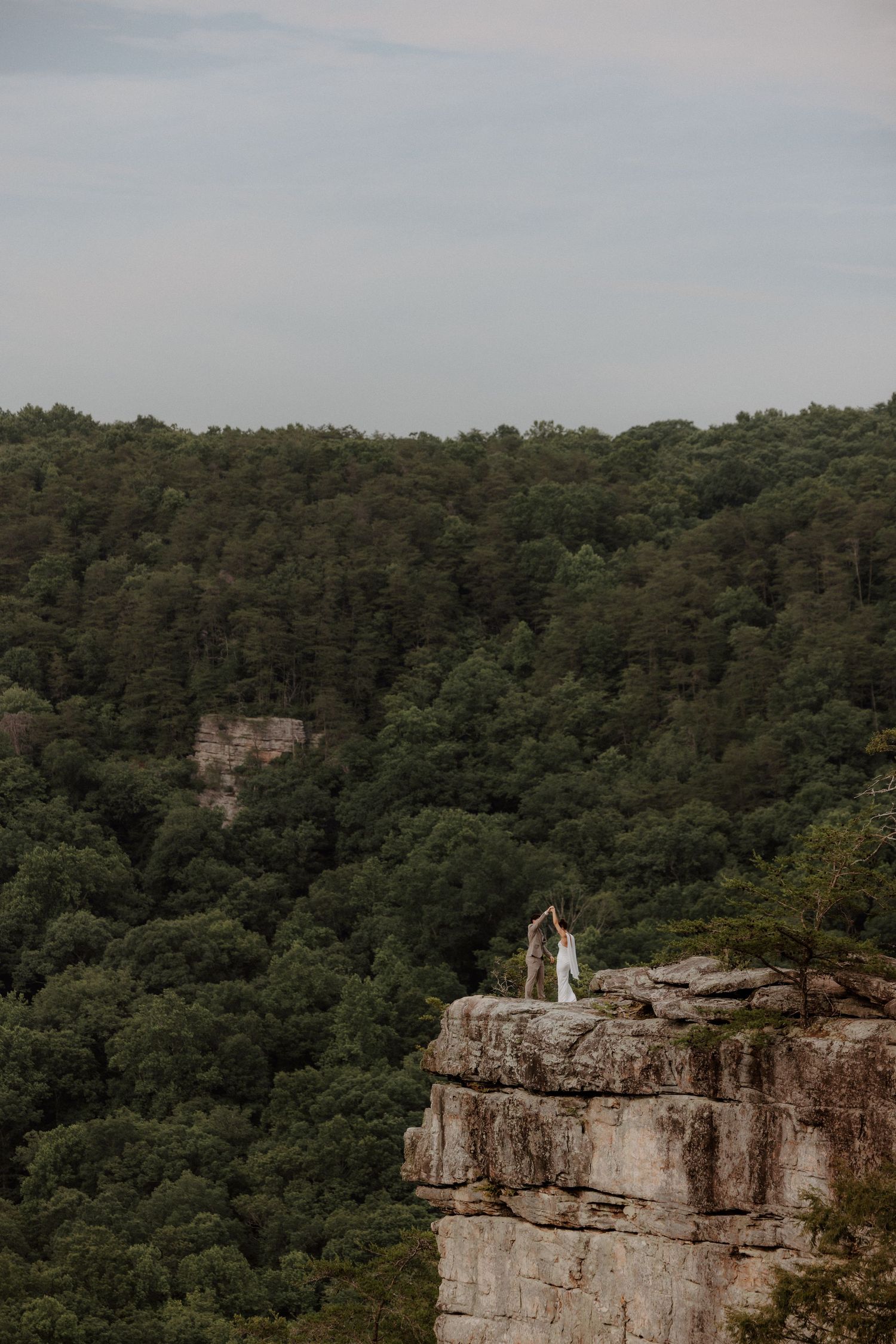 Bride and groom hold hands atop dramatic rocky cliff edge surrounded by dense green forest.