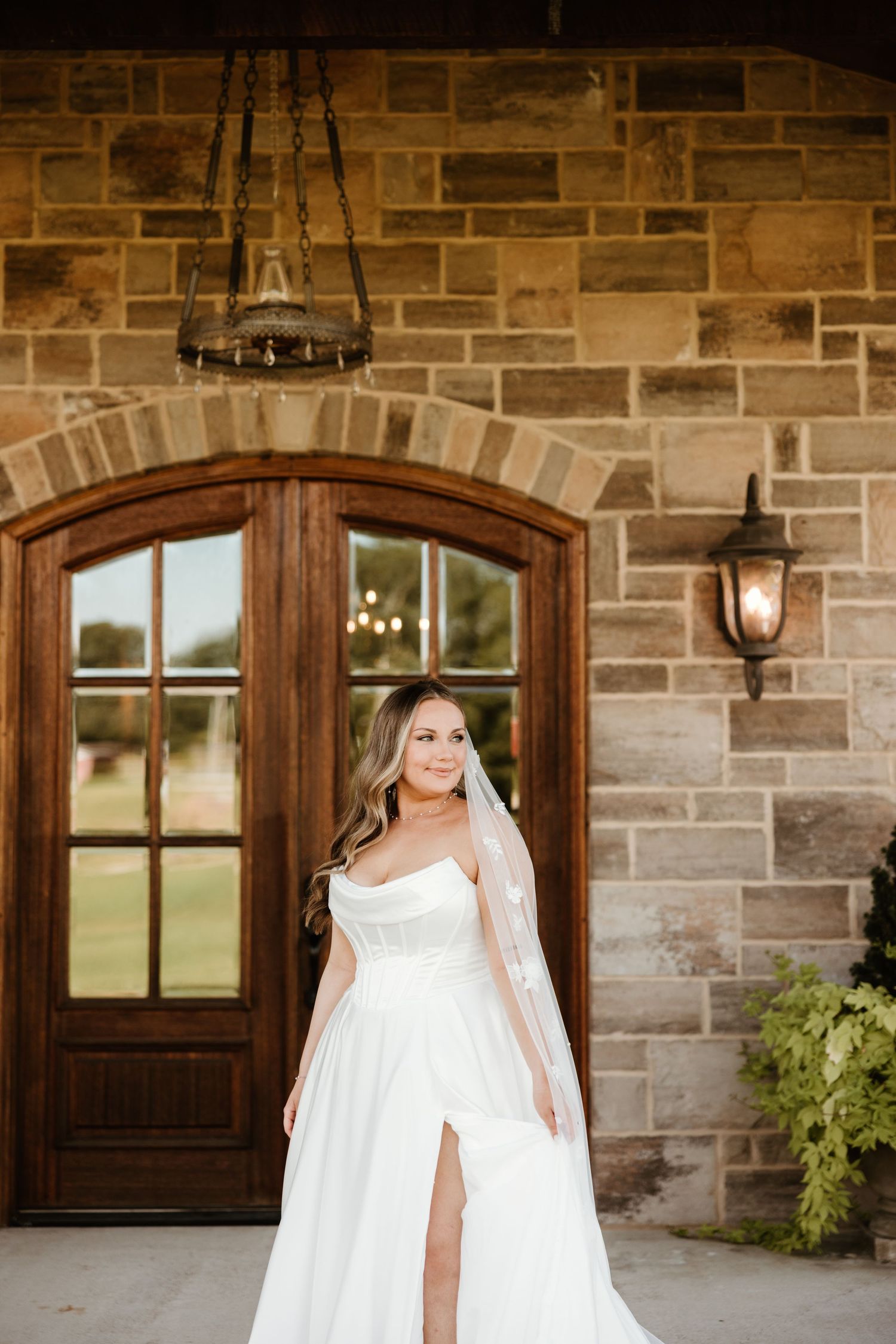Elegant white bridal gown with leg slit displayed in front of rustic stone wall and wooden arched doorway.