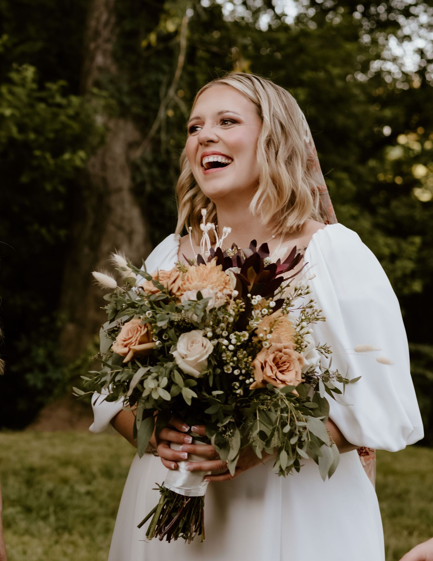 A laughing bride in a white dress holds a beautiful autumn-toned bouquet with peach roses and greenery in an outdoor setting.