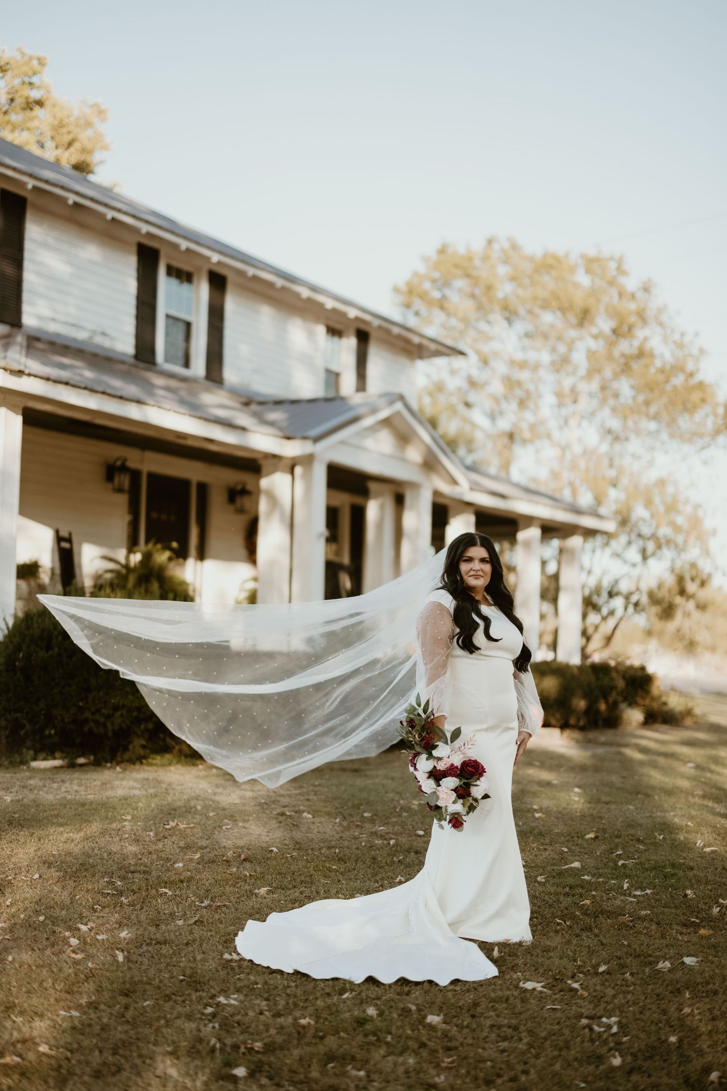 A bride in a flowing white dress with a dramatic long veil stands in front of a rustic white farmhouse in autumn.