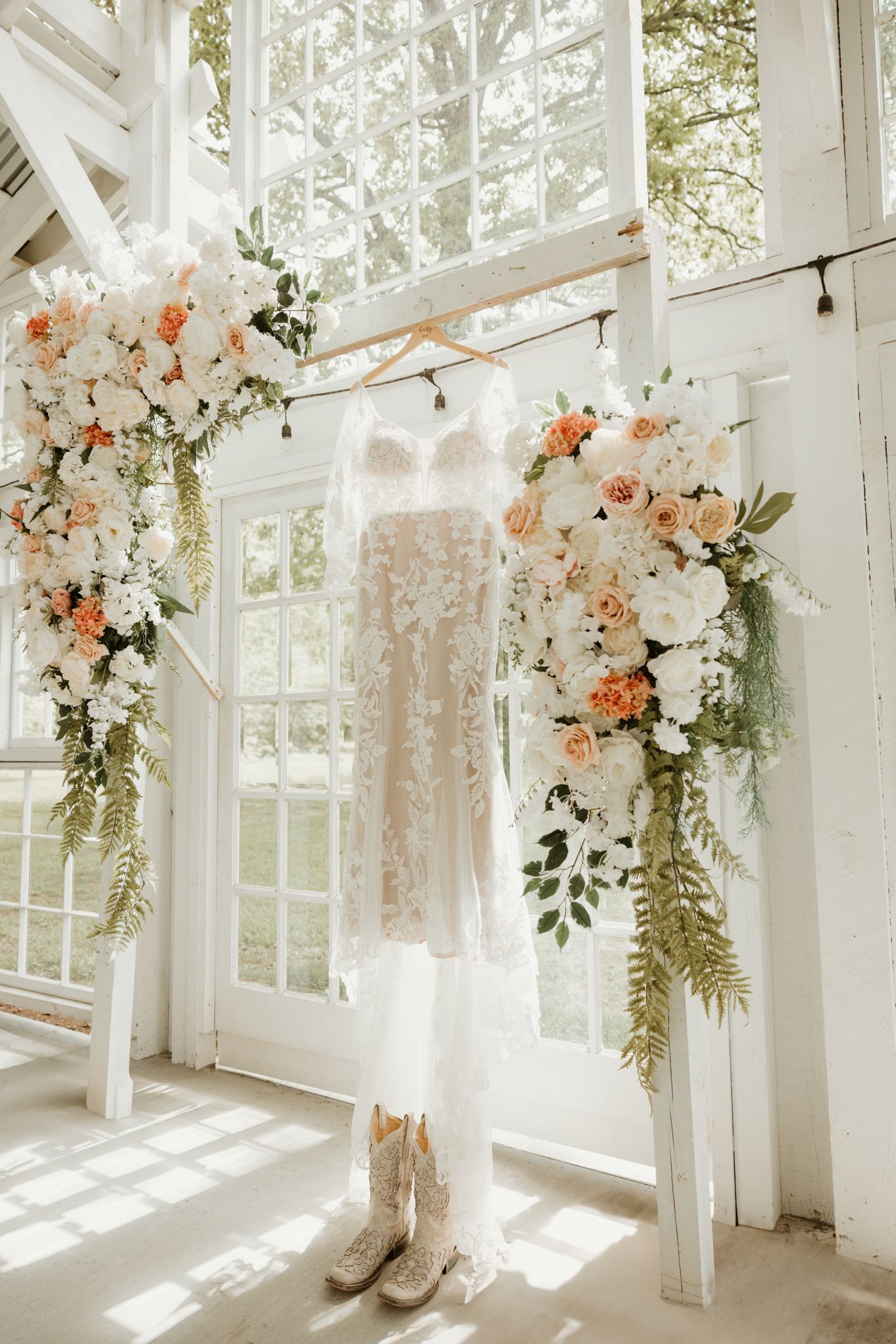A delicate lace wedding dress hangs between floral arrangements of peach and white blooms in a bright sunlit room.