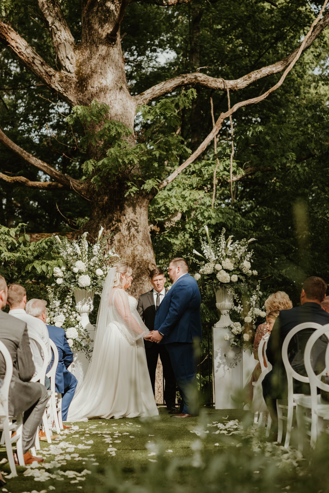 A wedding ceremony under a majestic tree with white floral arch and guests seated in elegant white chairs.