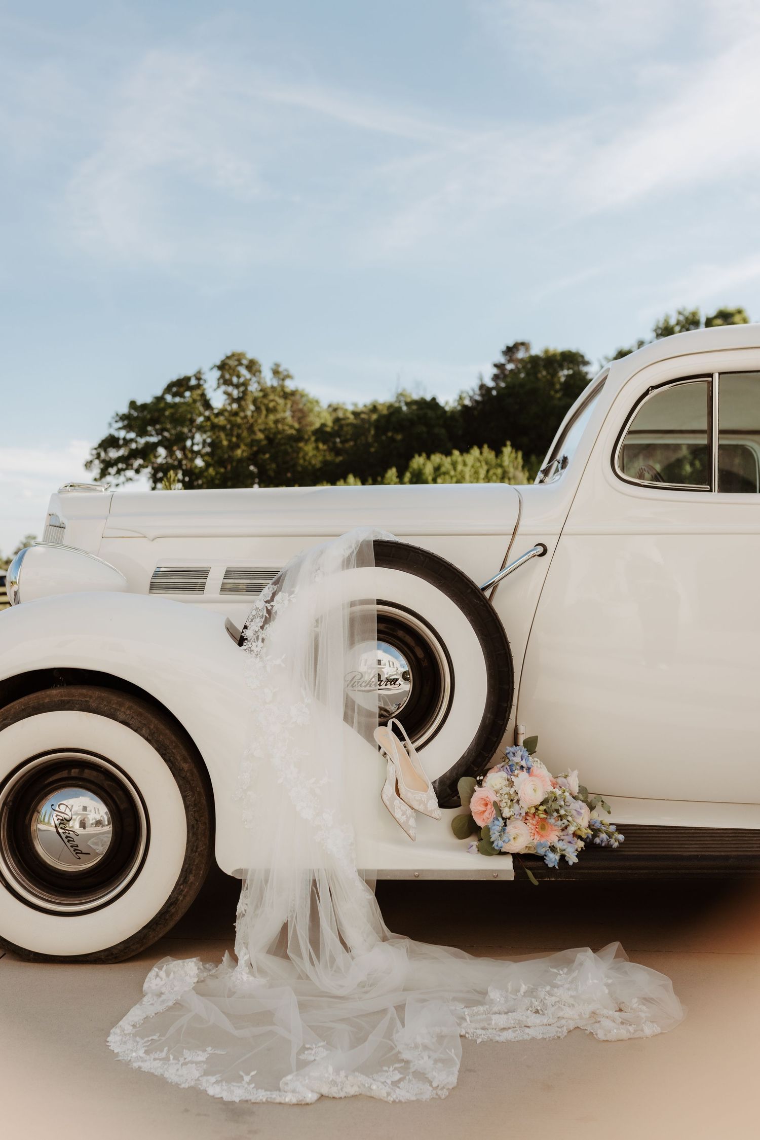 Vintage white classic car adorned with a bridal veil and floral bouquet outdoors.