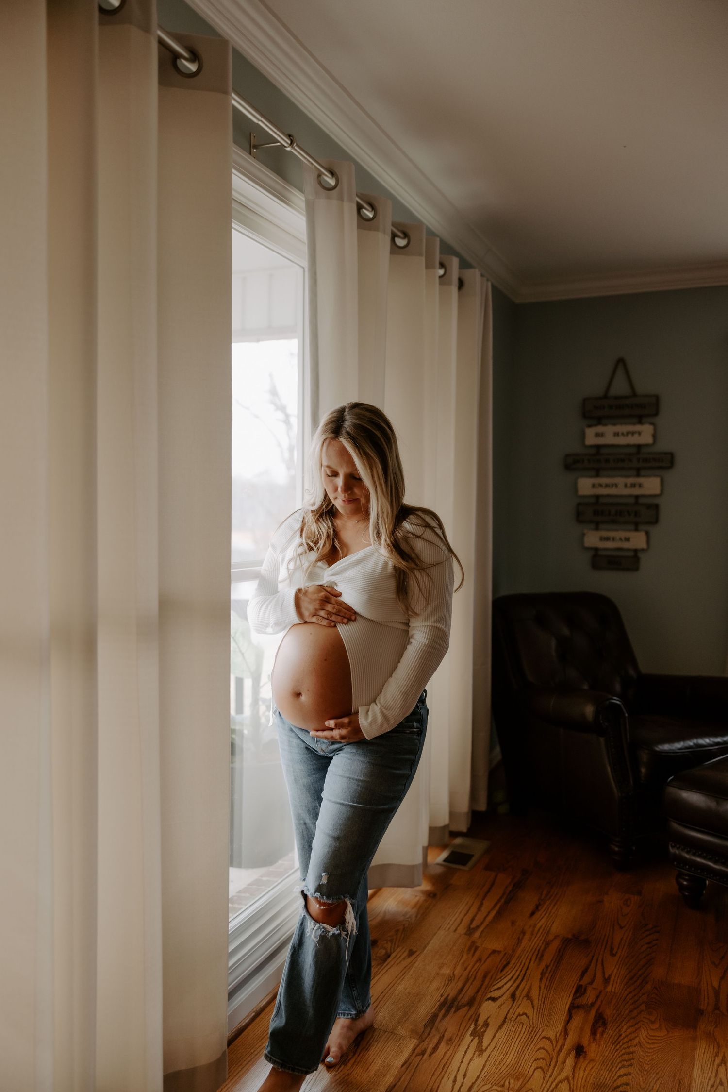 Pregnant woman in white top and ripped jeans cradling baby bump by a bright window in a cozy home.