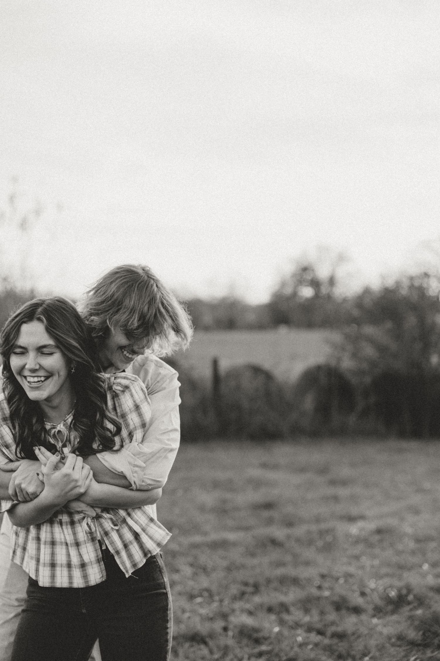 Black and white photo of a laughing woman being hugged from behind by a child in a grassy field.