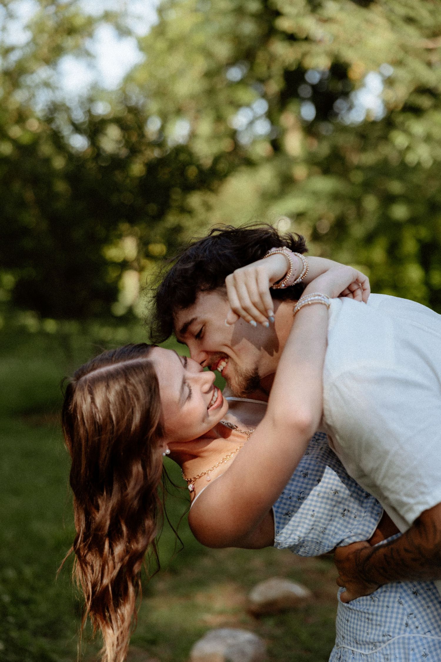 A joyful couple sharing a romantic dip kiss outdoors, surrounded by lush green trees in a sunlit park.