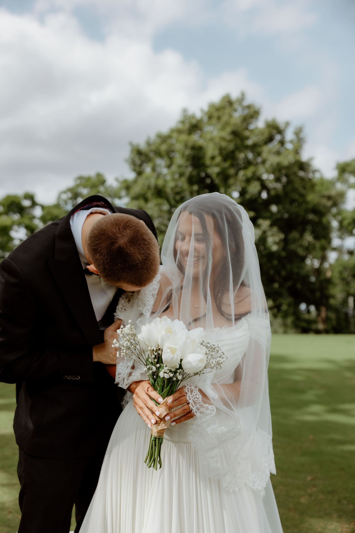 Bride in white gown and veil holds bouquet while groom in black suit leans in for a kiss outdoors.