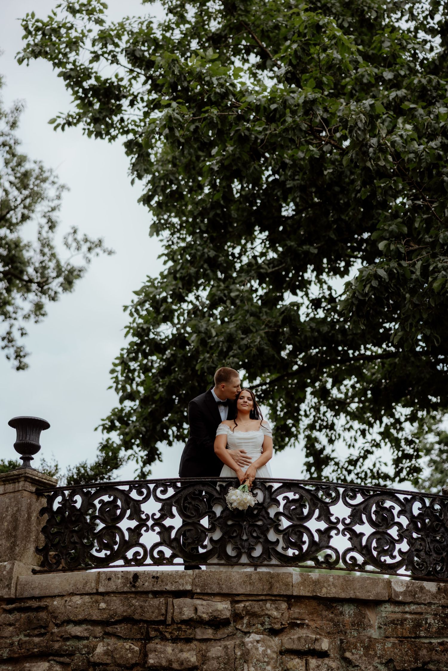 Bride and groom share a romantic moment on an ornate iron balcony surrounded by lush green trees.