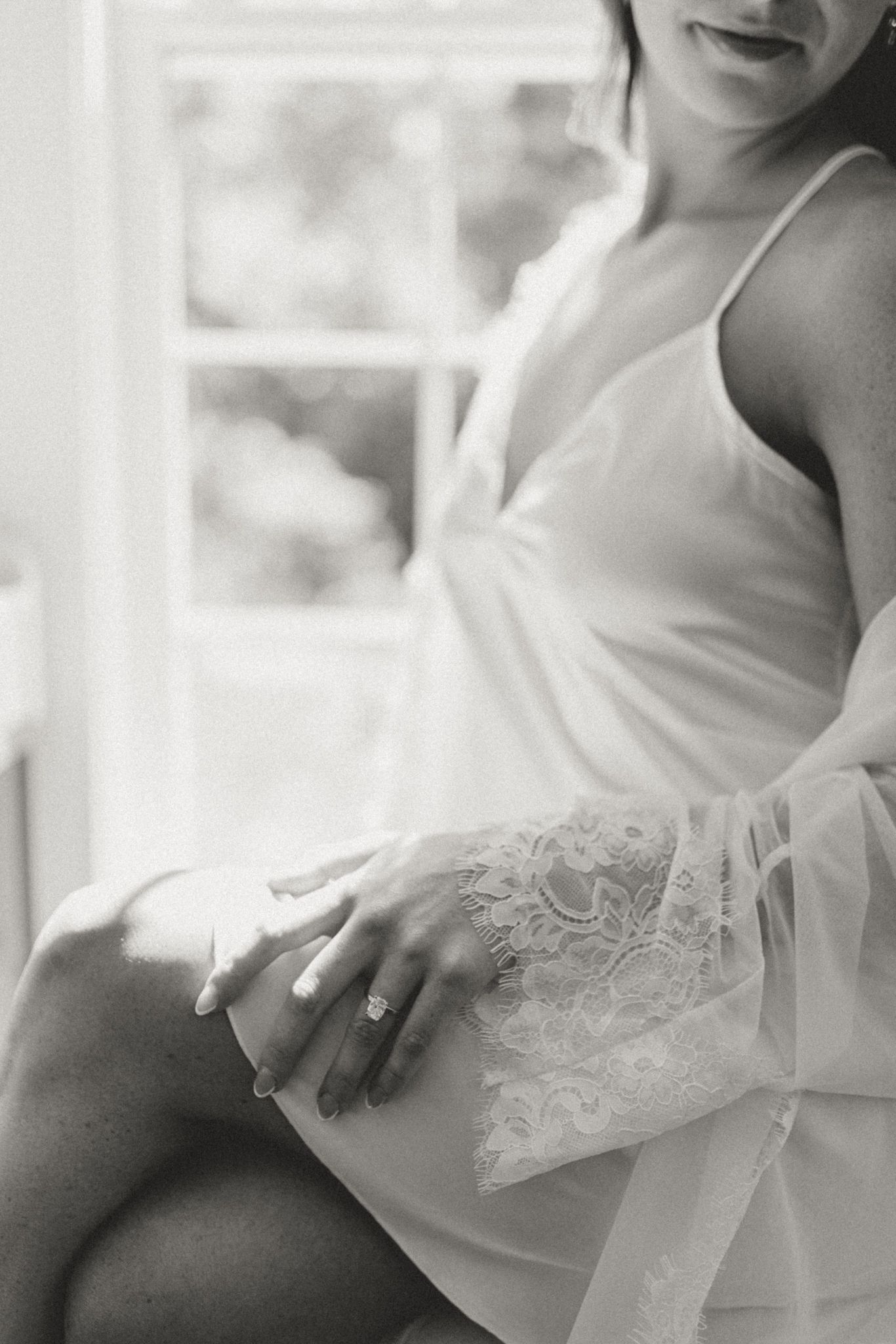 Black and white close-up of a delicate white lace dress detail and hand near a sunlit window.