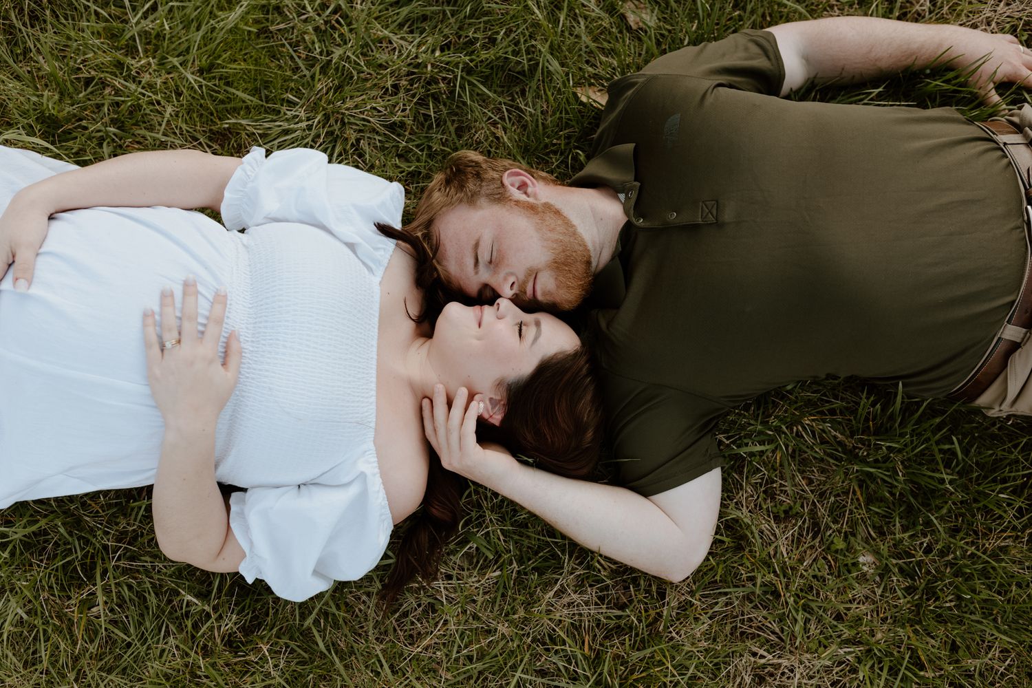 A couple lies on grass, heads together, wearing white and green clothing in a romantic outdoor scene.