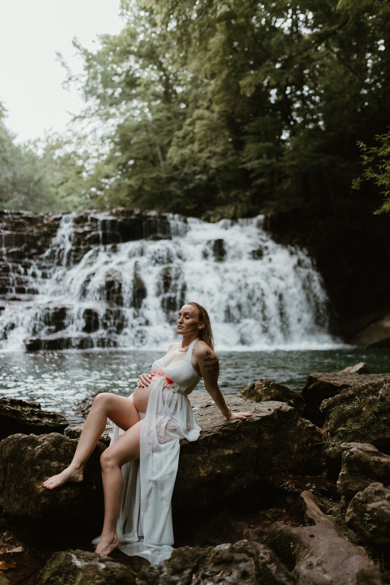 A dreamy maternity photoshoot features a flowing white dress against a cascading waterfall backdrop in nature.