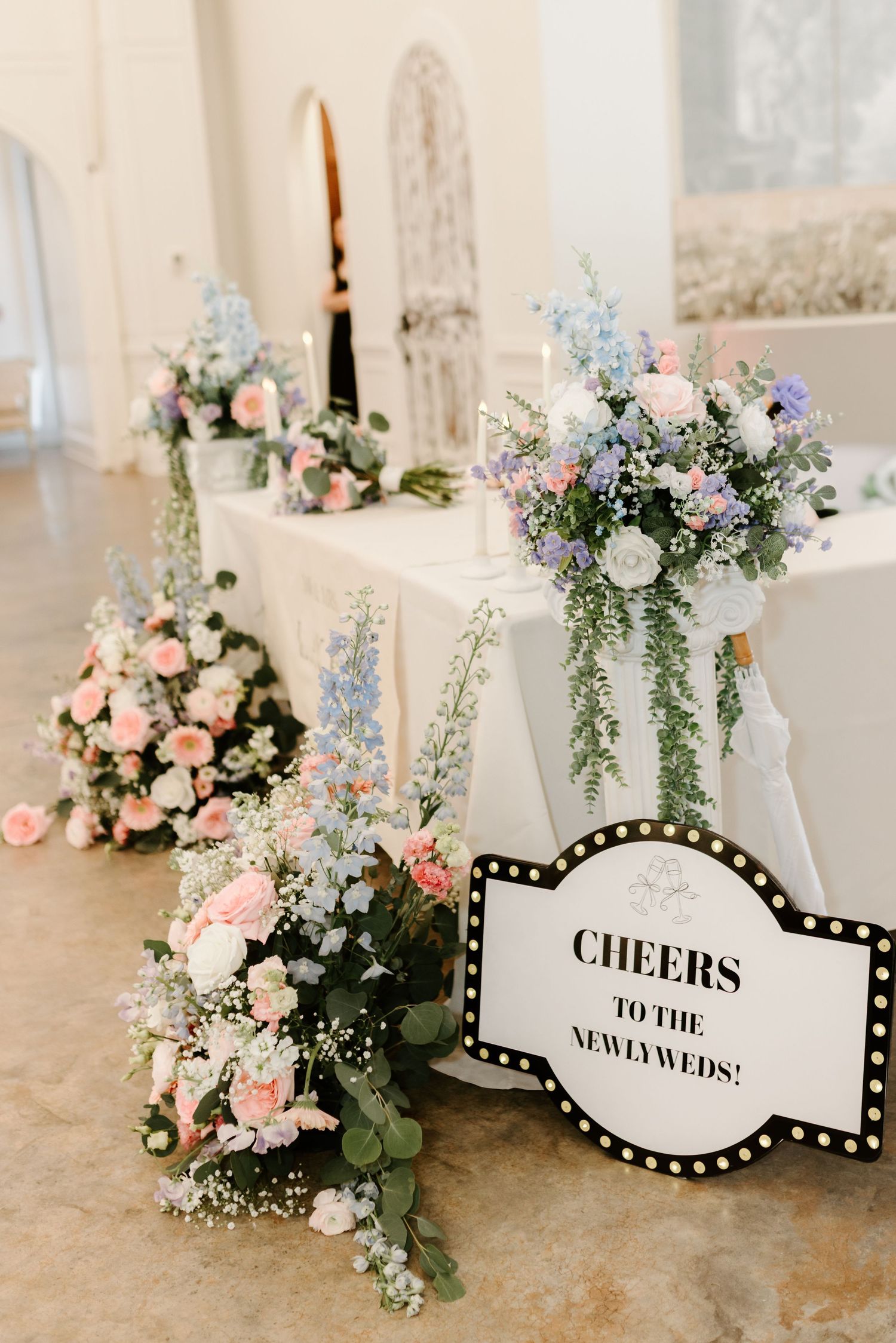 Elegant wedding sweetheart table adorned with pink, white, and blue floral arrangements and a marquee sign reading Cheers to the Newlyweds.