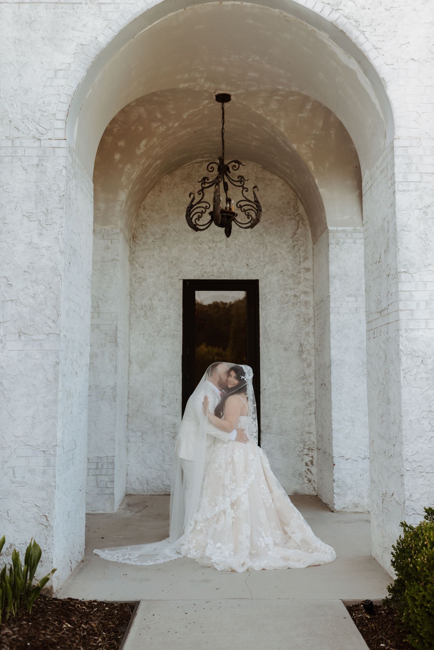 Bride in lace gown and veil stands under white stone arch with chandelier at wedding venue.