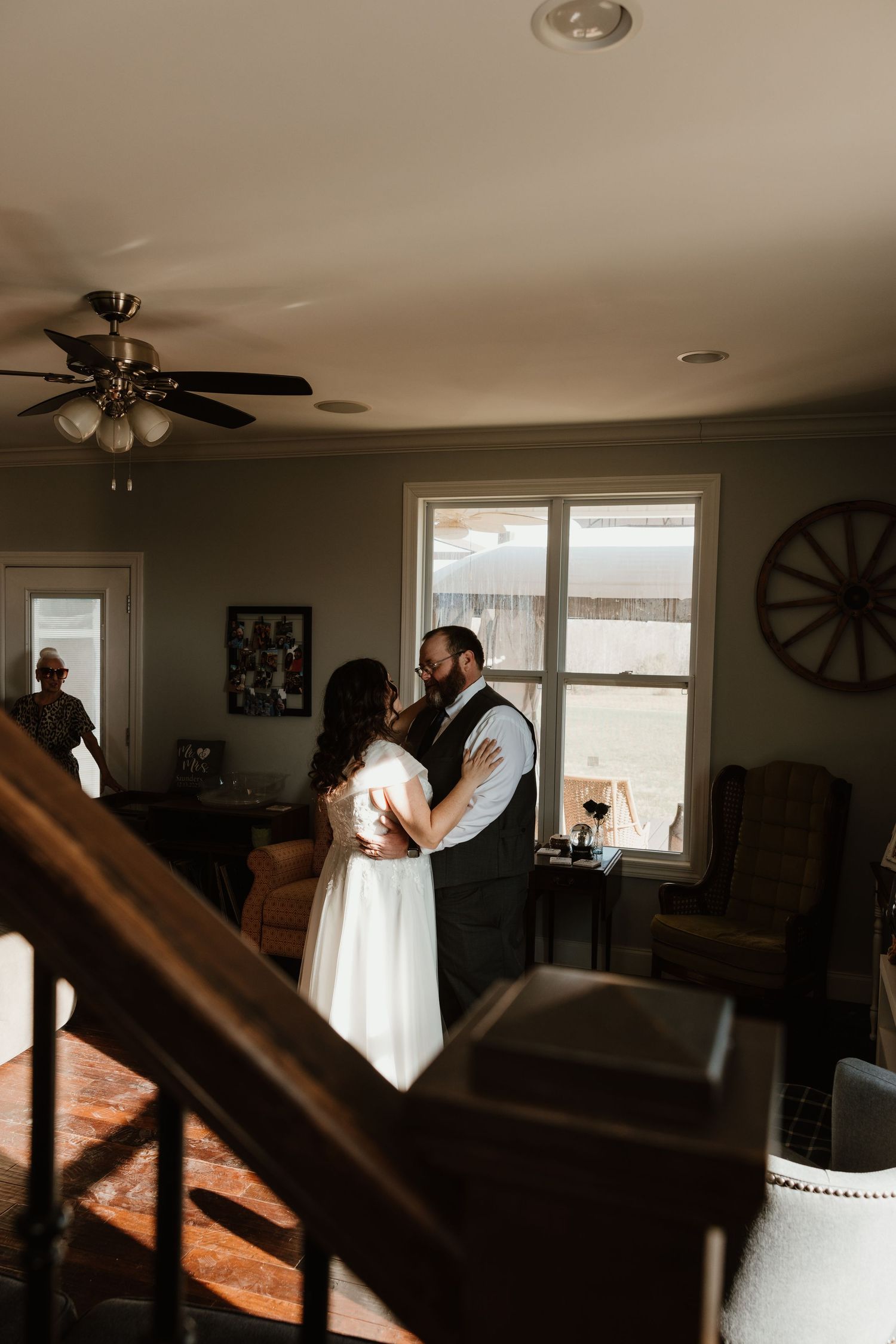 A bride and groom share a tender embrace near a sunlit window in a rustic farmhouse living room on their wedding day.