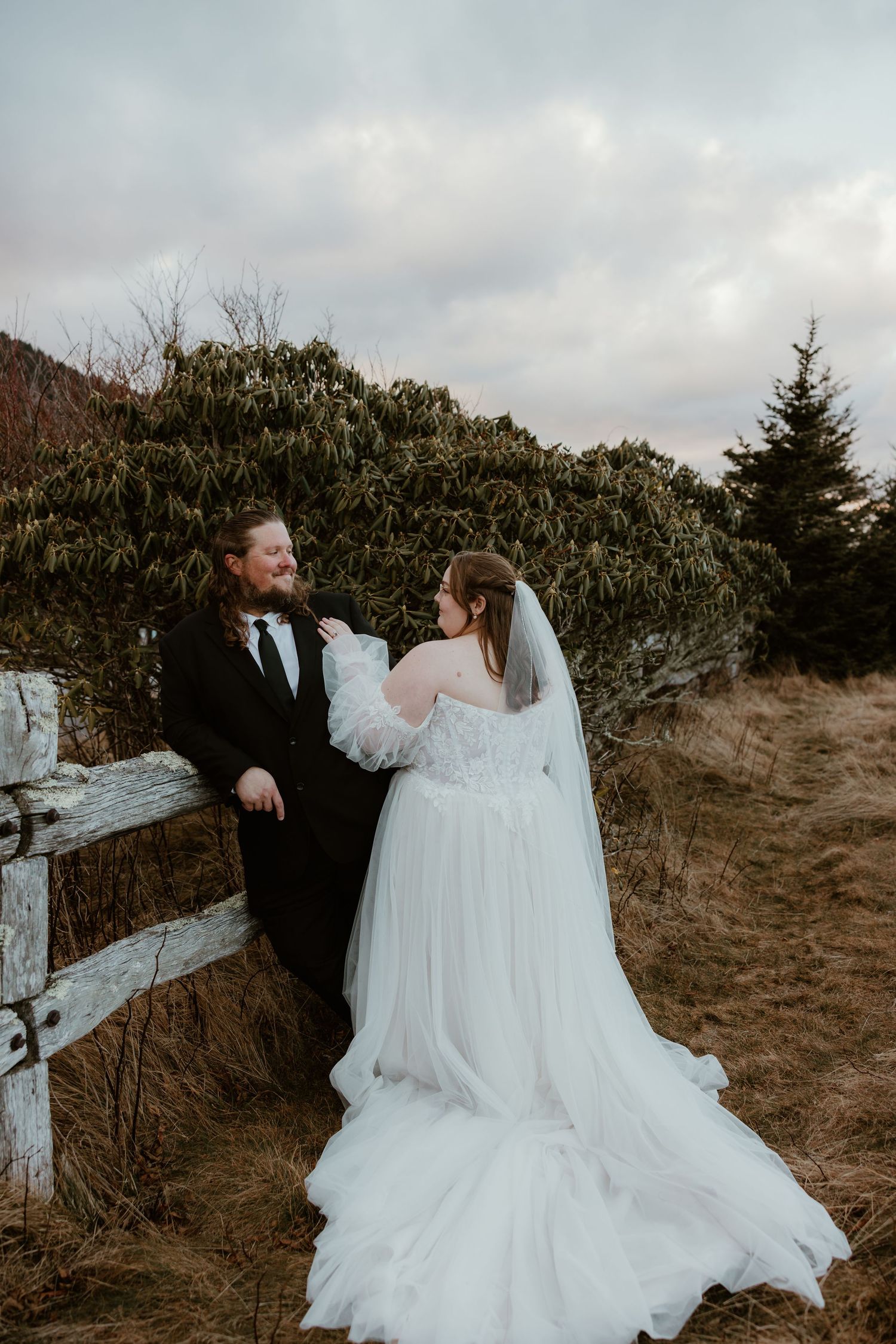 Bride in white tulle gown and groom in black suit share a joyful moment by rustic fence with mountain greenery.