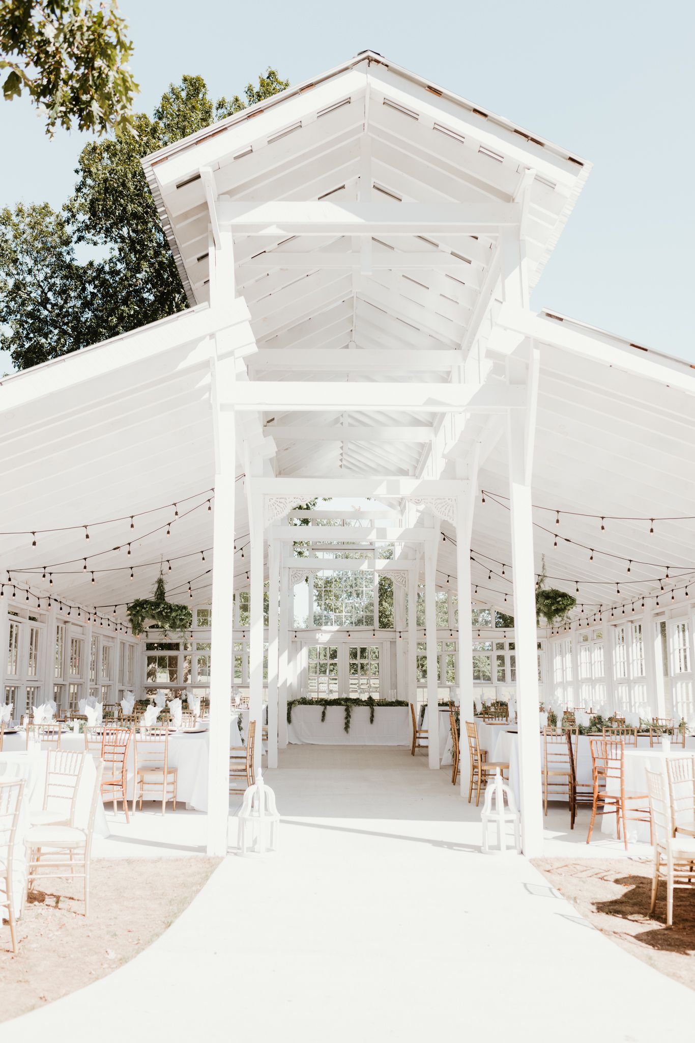 A bright white outdoor wedding pavilion with string lights and wooden chairs set up for an elegant ceremony or reception.