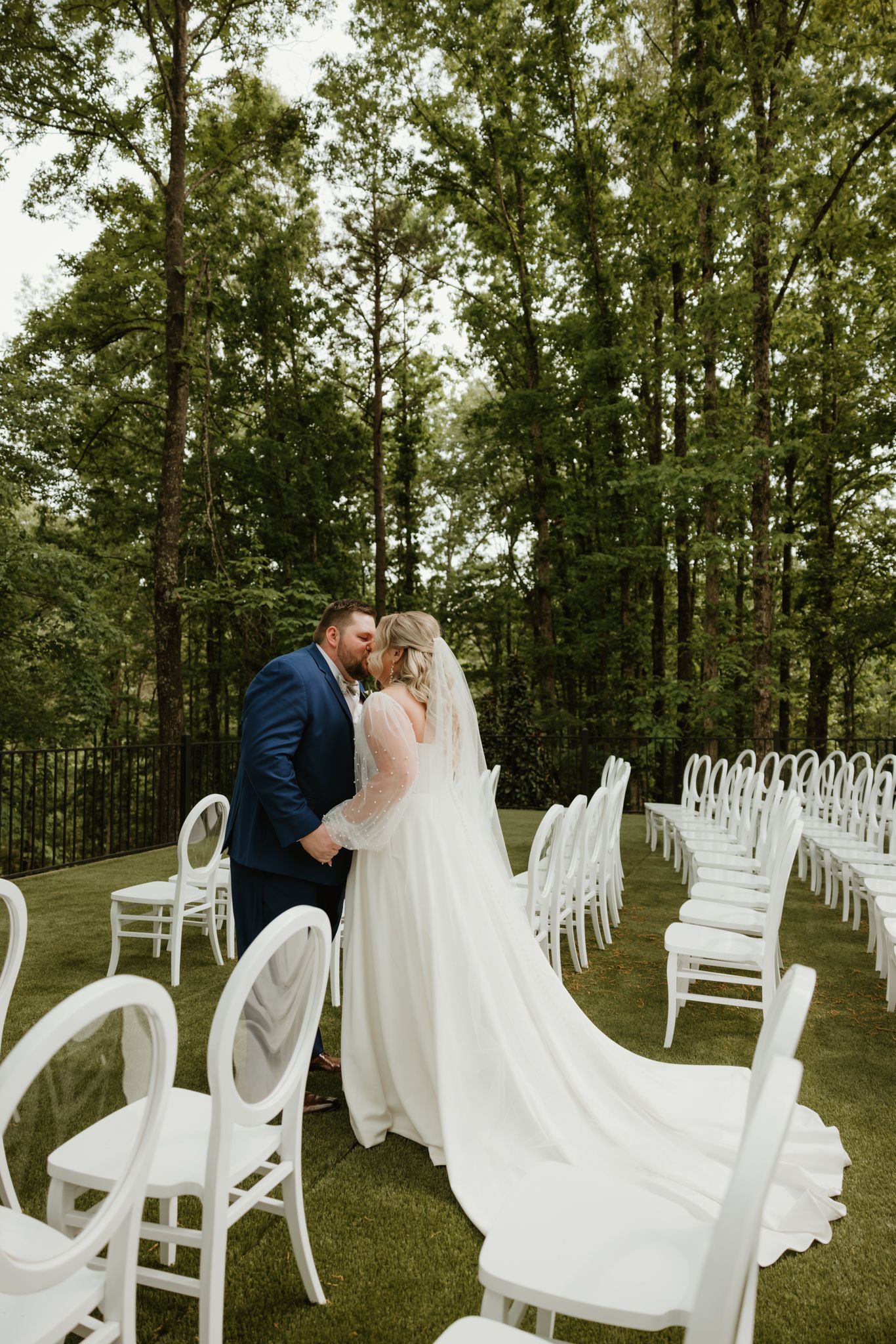 White wedding chairs arranged in rows on a green lawn with tall trees creating a natural backdrop.