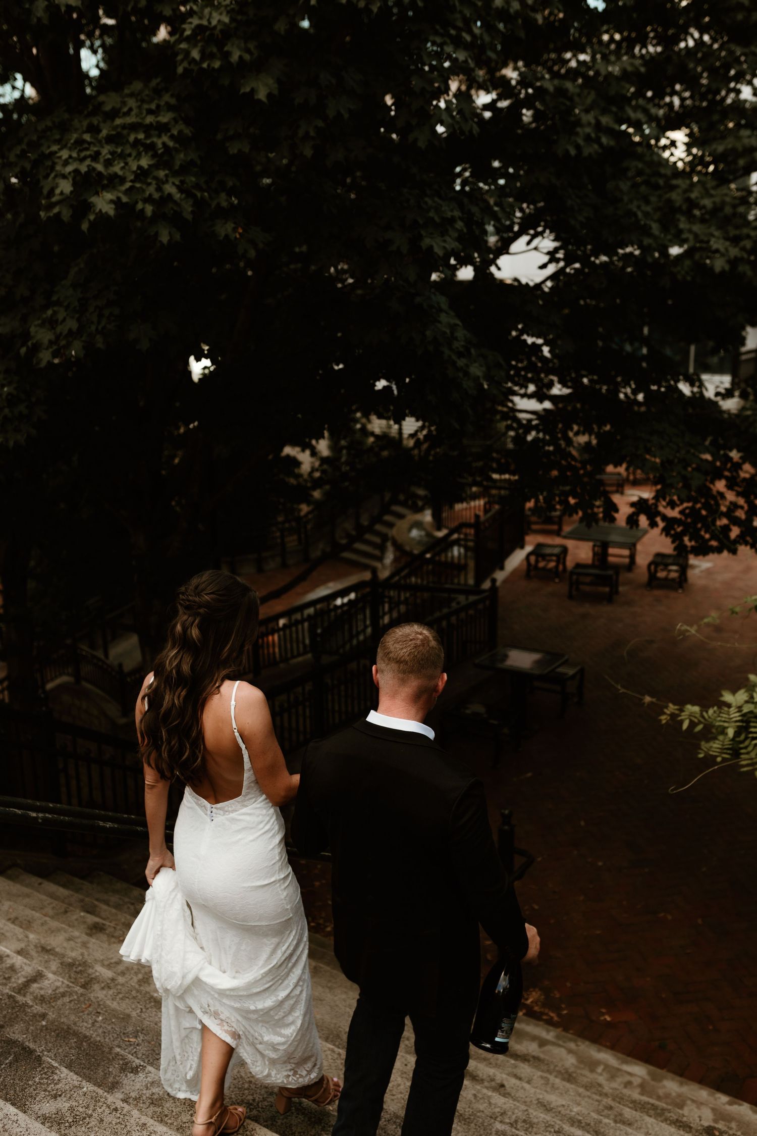 Couple in wedding attire walking down stone steps overlooking an outdoor patio area with tables at dusk.