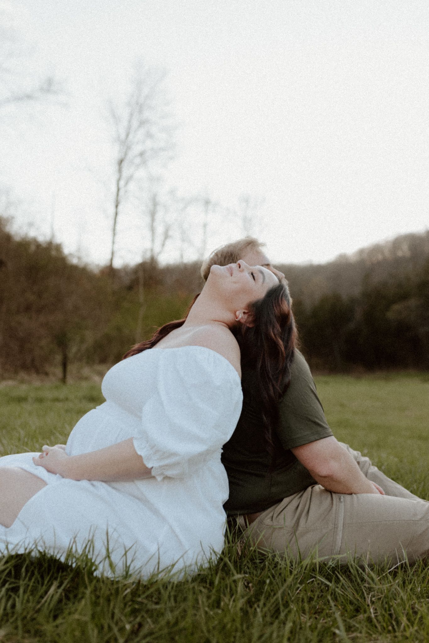 A maternity photoshoot features a couple sitting together in a grassy field at sunset.