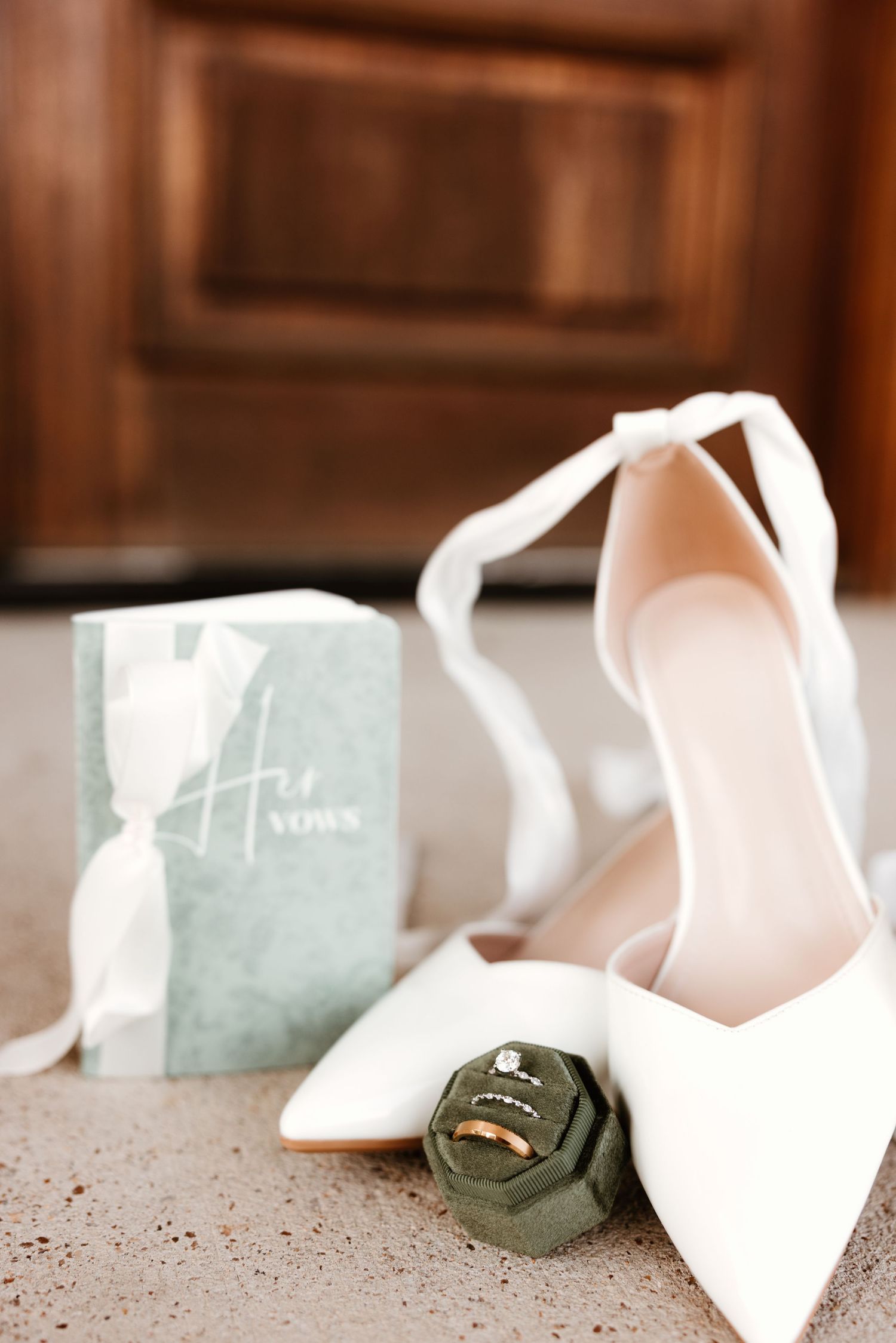 Elegant white wedding shoes displayed alongside wedding rings on moss green velvet ring box and light blue invitation.