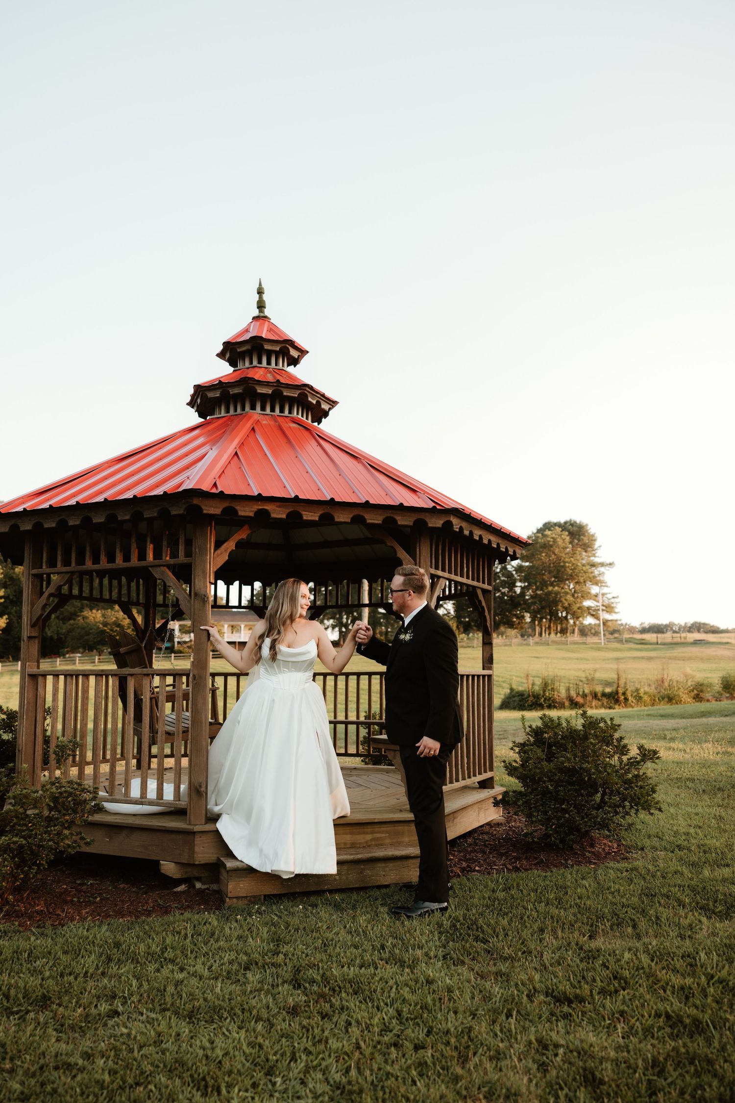 A couple shares a romantic moment at sunset in a red-roofed wooden gazebo surrounded by manicured lawn.