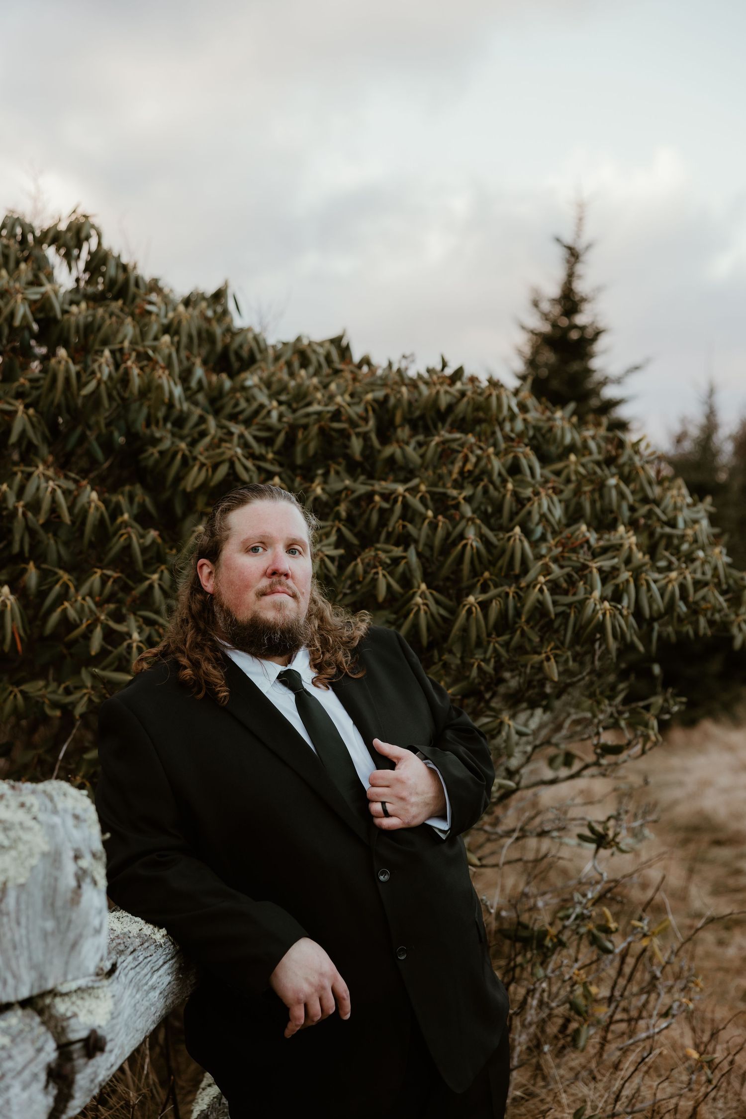 Groom with long hair in black suit leans against log near evergreen shrubs under cloudy sky at mountain wedding.