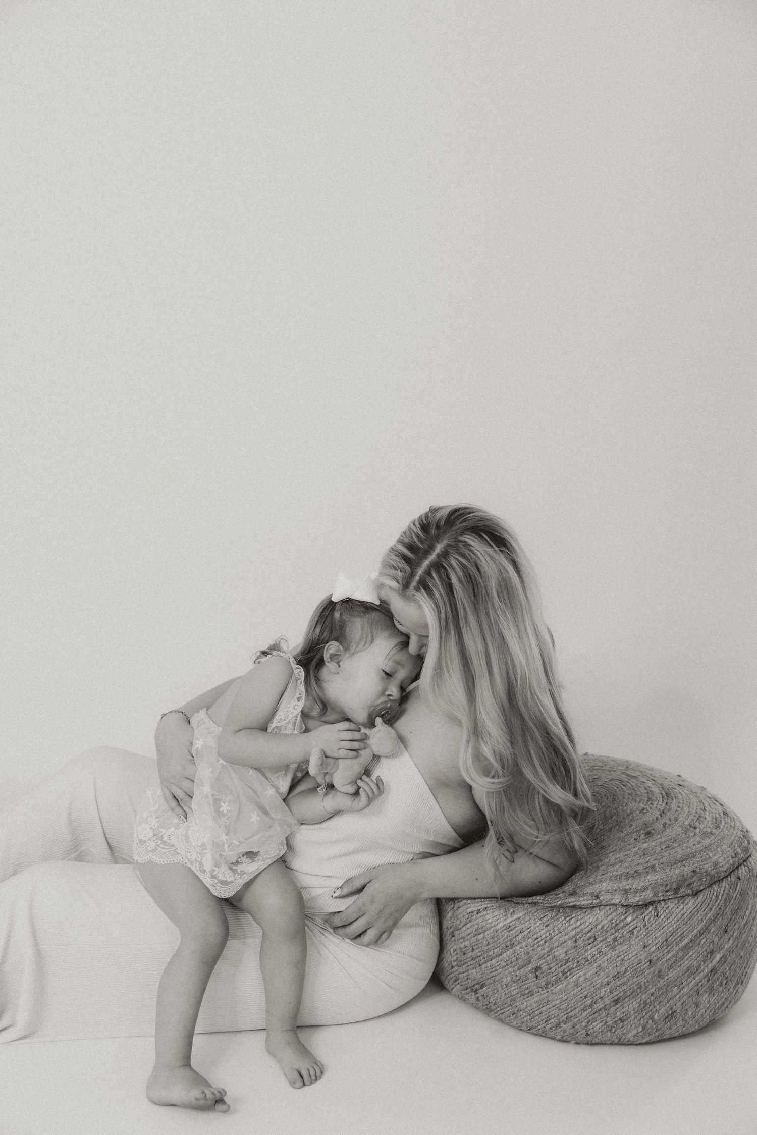 Black and white photo of a mother tenderly holding her baby girl on a white pouf in a bright studio setting.