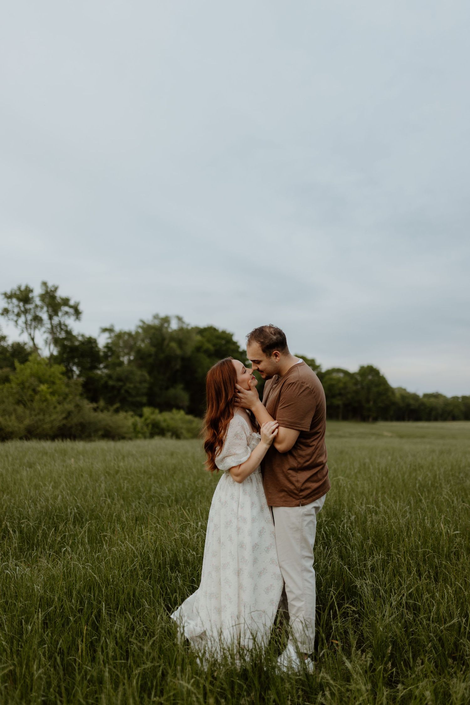 A couple shares a romantic moment kissing in a lush green field, woman in white dress, man in brown shirt.
