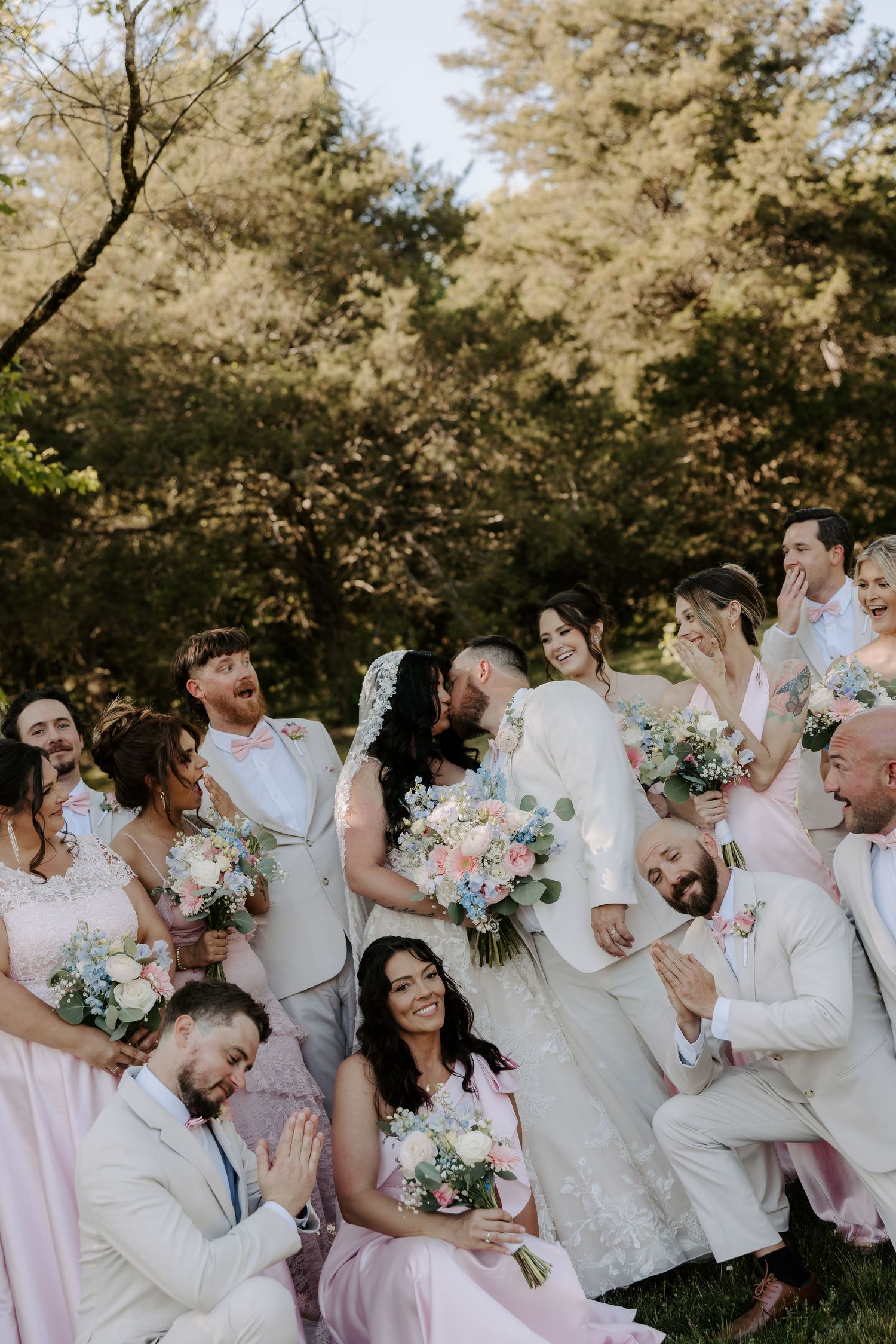 Wedding party laughing and celebrating outdoors with bride and groom kissing, bridesmaids in pink dresses holding floral bouquets.