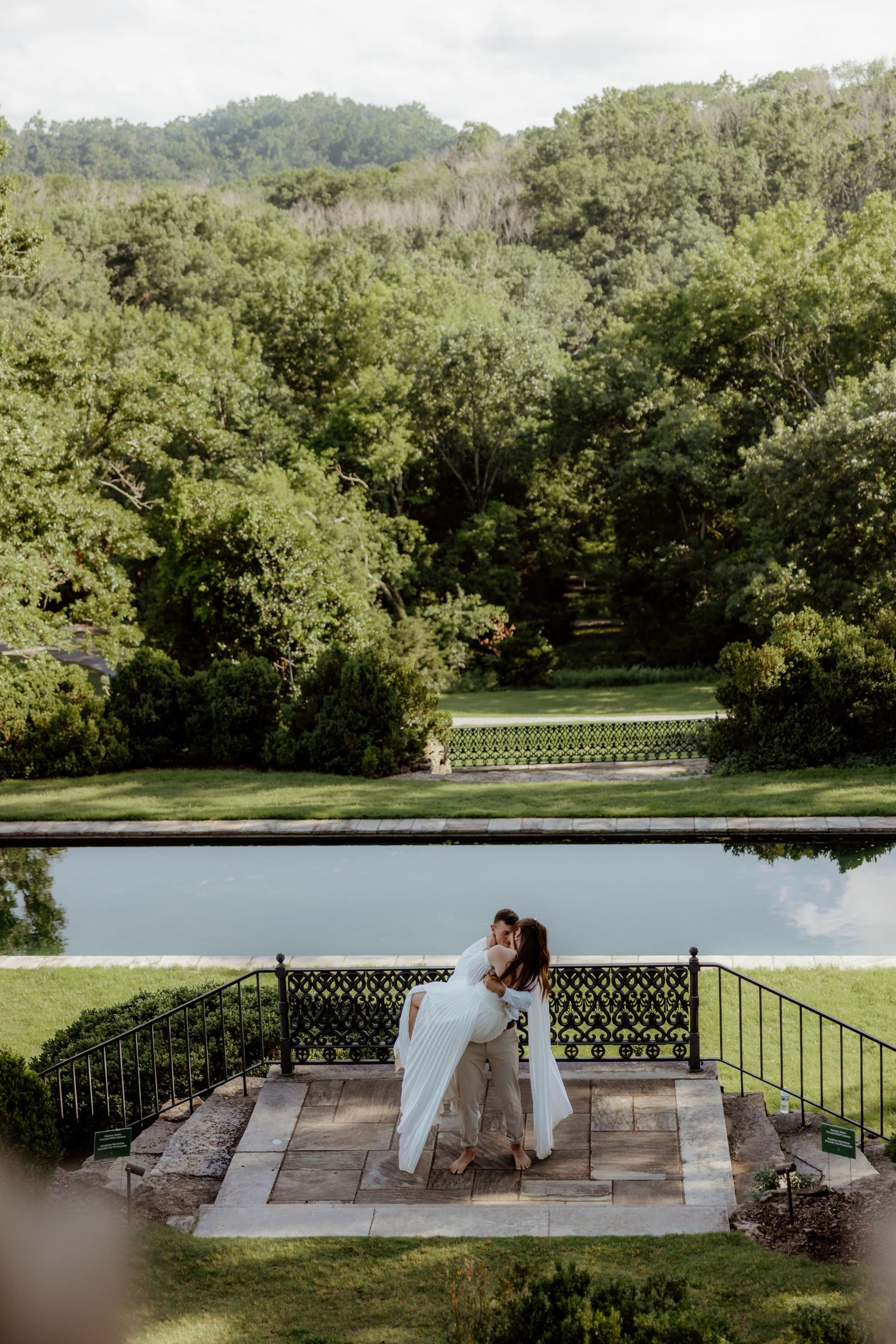 Bride and groom embrace on a stone terrace beside a reflecting pool surrounded by lush green gardens.