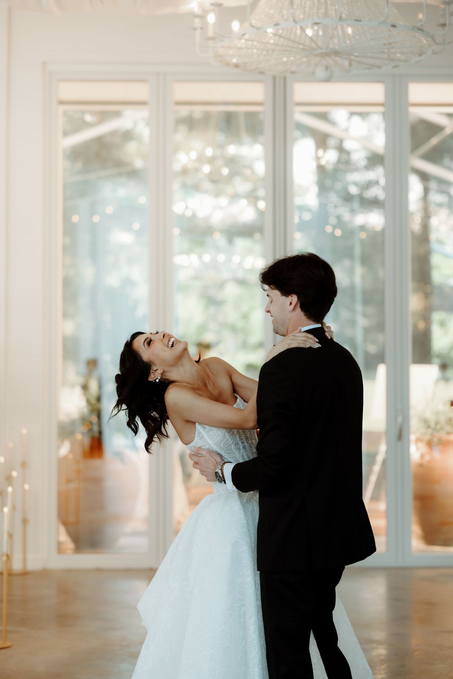 Bride and groom share their first dance in a bright white ballroom adorned with candles and chandelier.