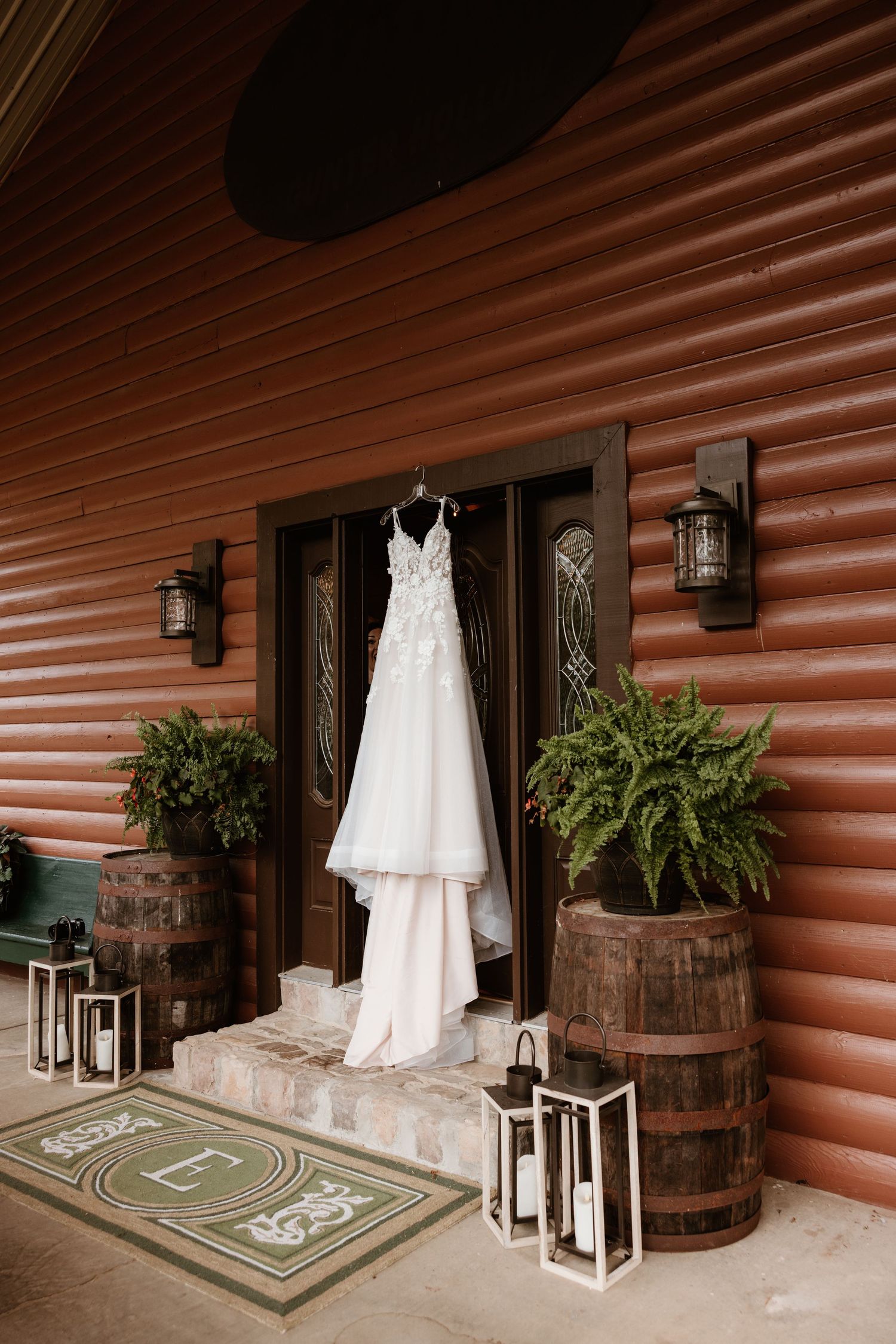 White wedding dress displayed on a log cabin porch with rustic wooden barrels and decorative lanterns creating an elegant setting.