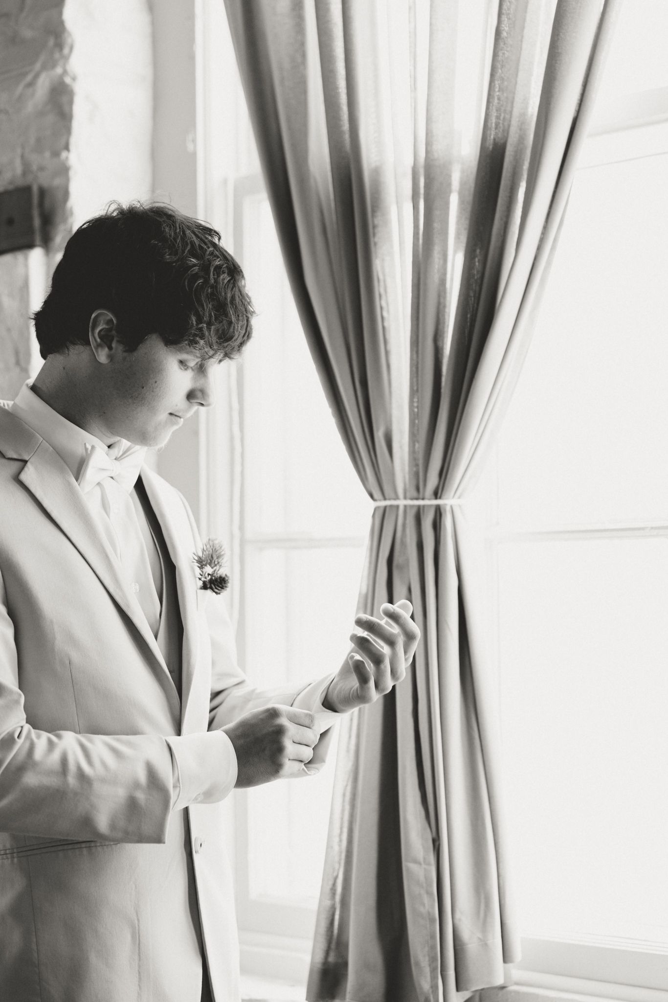 Black and white photo of a groom in a light suit checking his phone by a curtained window.