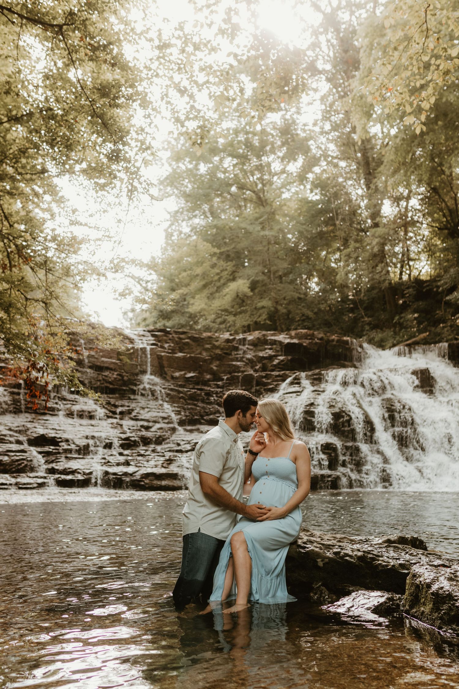 A couple stands in shallow waterfall during maternity photoshoot wearing casual attire.